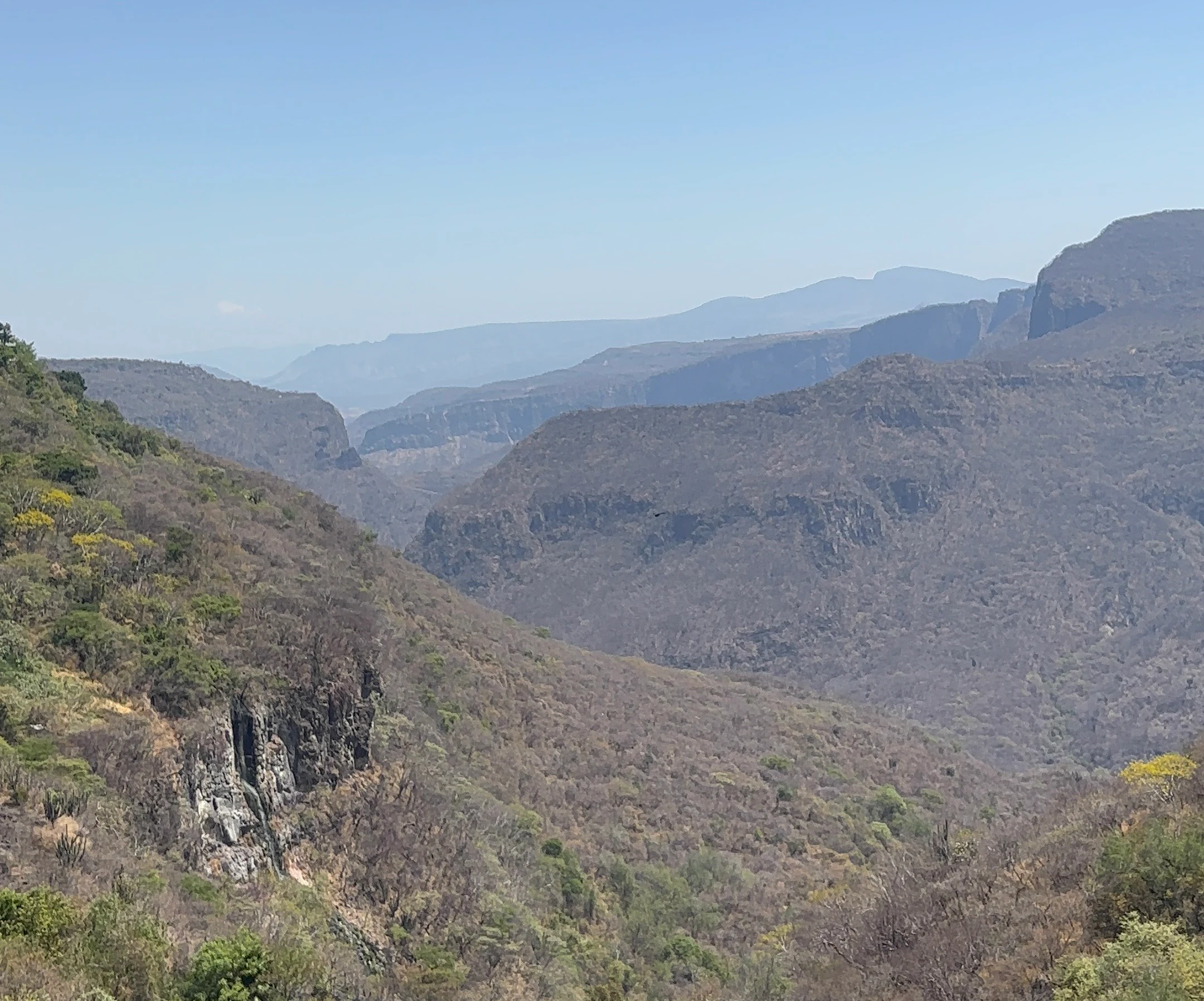 A wide scenic shot of a dry canyon landscape with steep rocky slopes covered in sparse vegetation under a hazy blue sky.