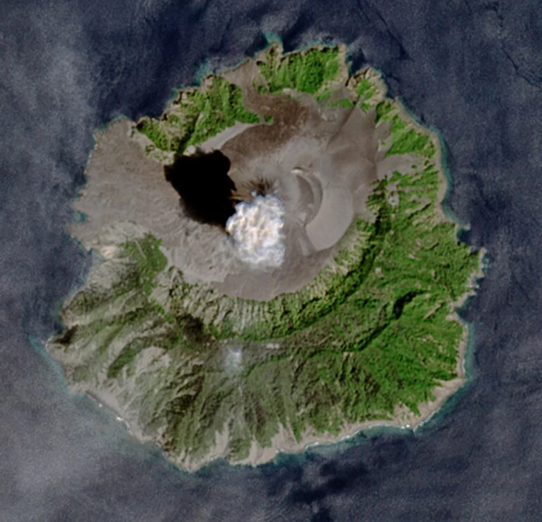 An aerial satellite image of Barren Island in the Andaman Sea showing the circular volcanic island with a central crater emitting a plume of smoke.