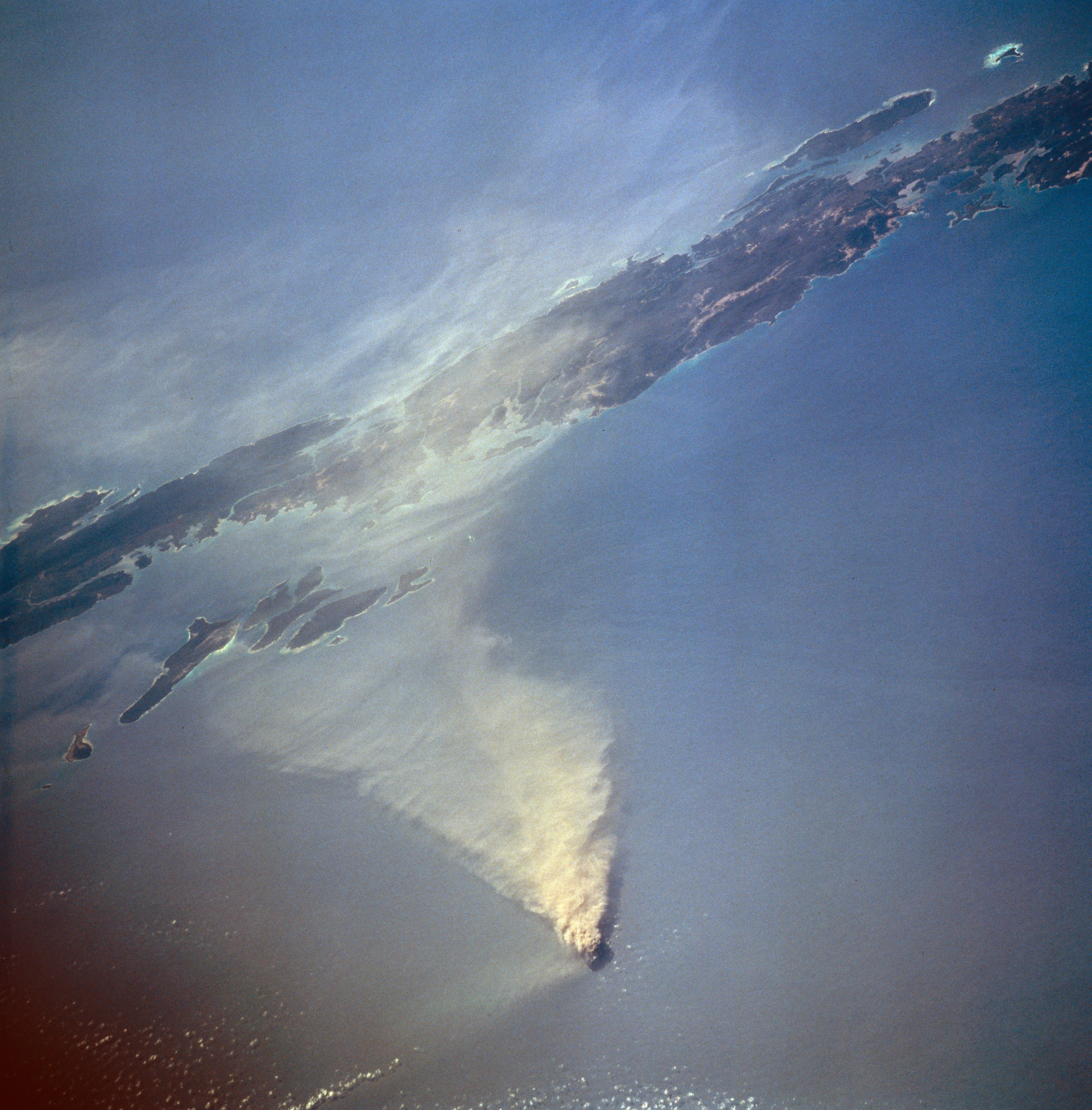 High angle aerial satellite view of a small volcanic island in the ocean with a large plume of smoke and ash extending across the water surface.