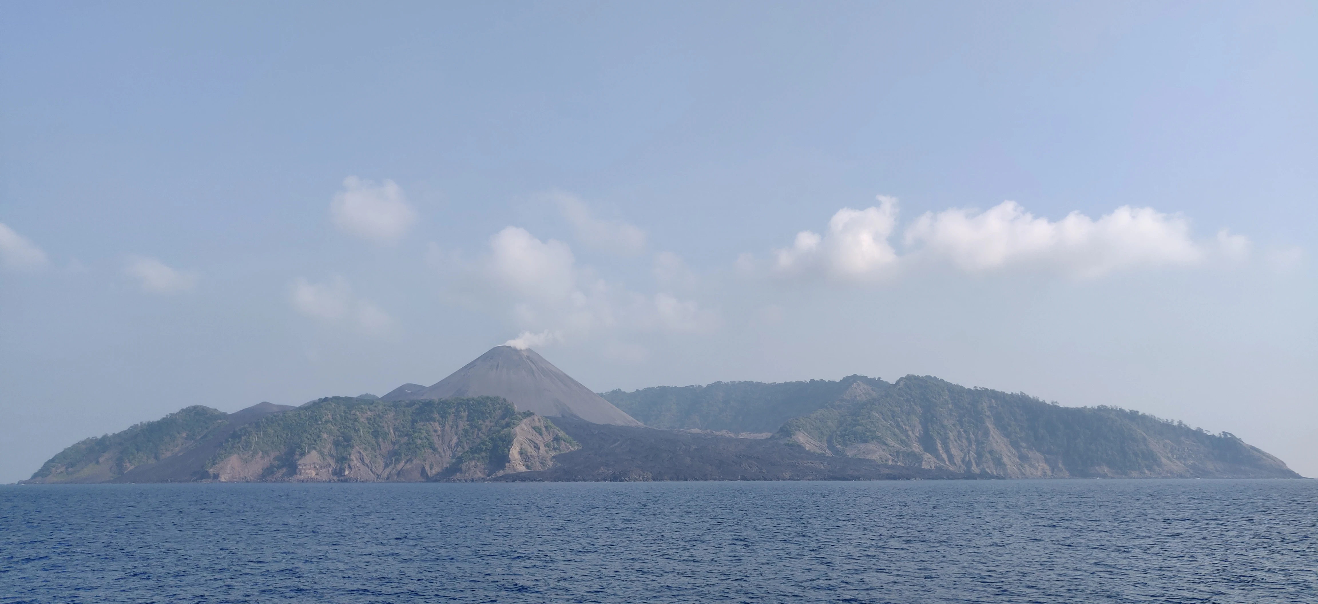 A wide shot of a volcanic island rising from the sea, with a conical peak emitting a faint plume of smoke and rugged, partially vegetated slopes.