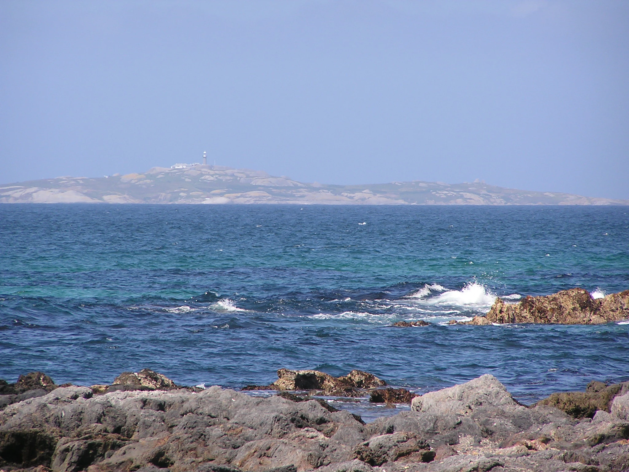 A distant view of a low-lying island across dark blue ocean waters, viewed from a rocky shoreline in the foreground.