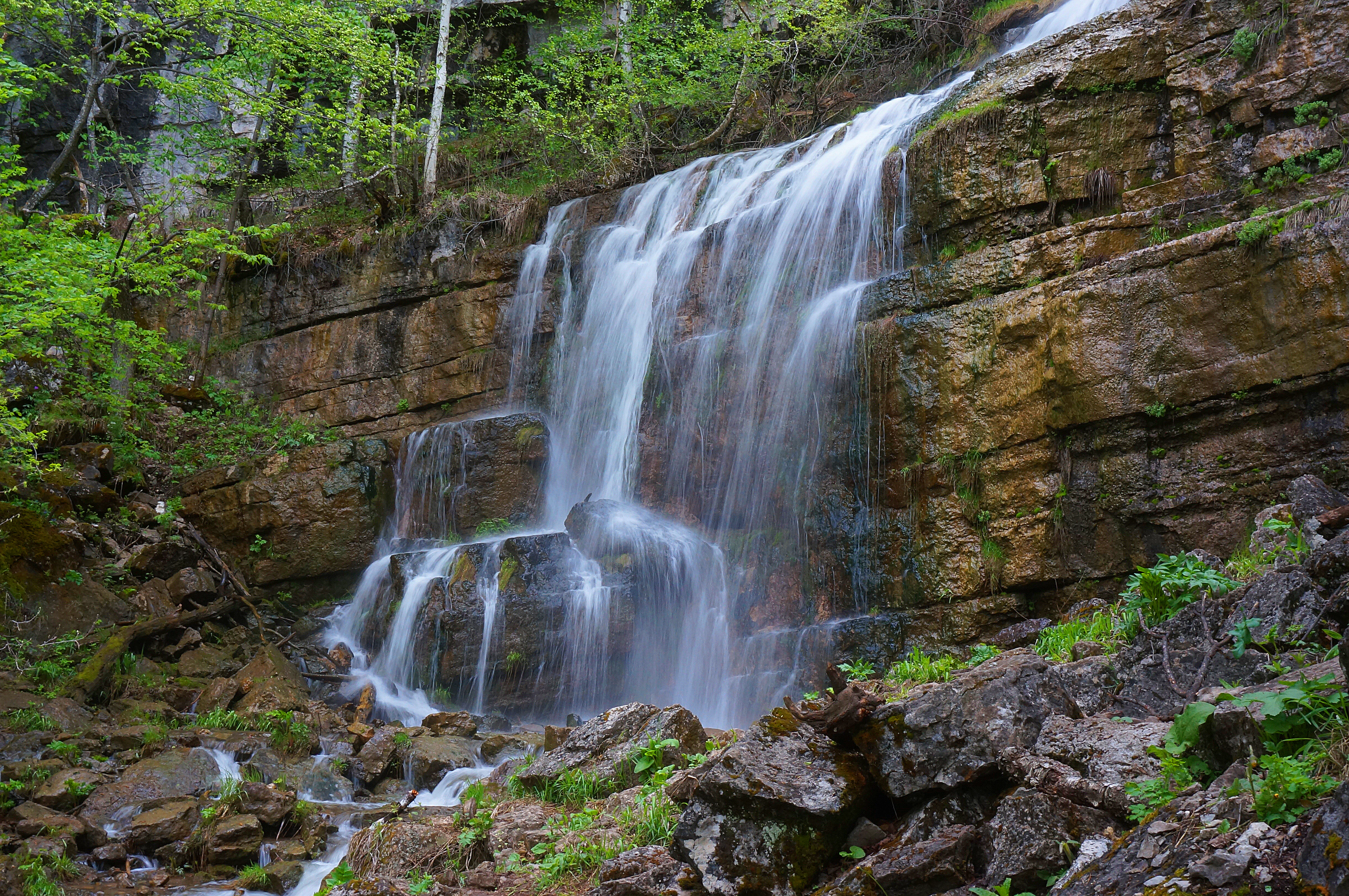 Una cascada de varios niveles fluye sobre capas horizontales y erosionadas de roca caliza marrón, rodeada de frondosa vegetación forestal verde.