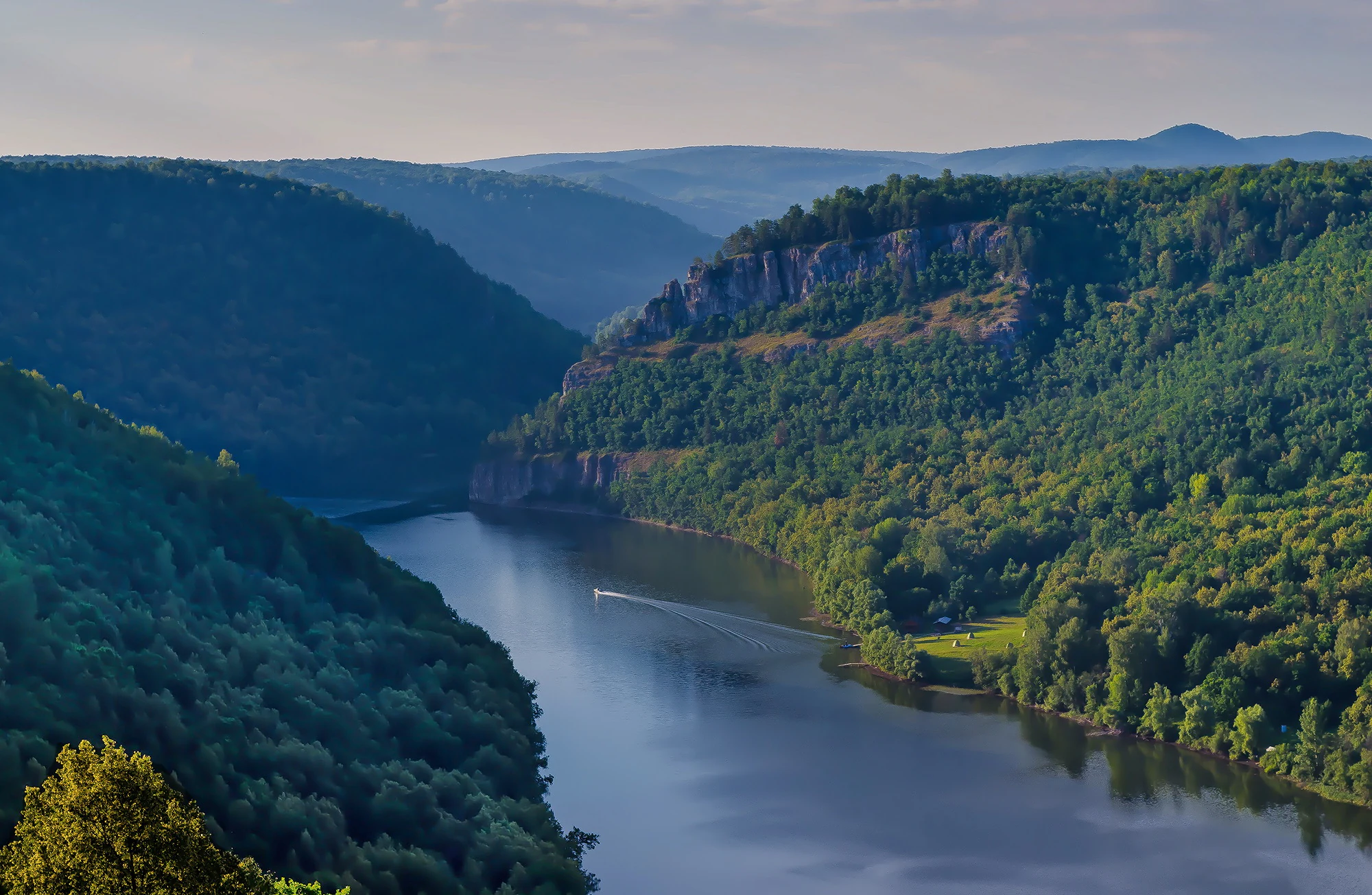 Wide river flowing through a lush green valley with forested hills on both sides and a prominent cliff face visible on the right hillside