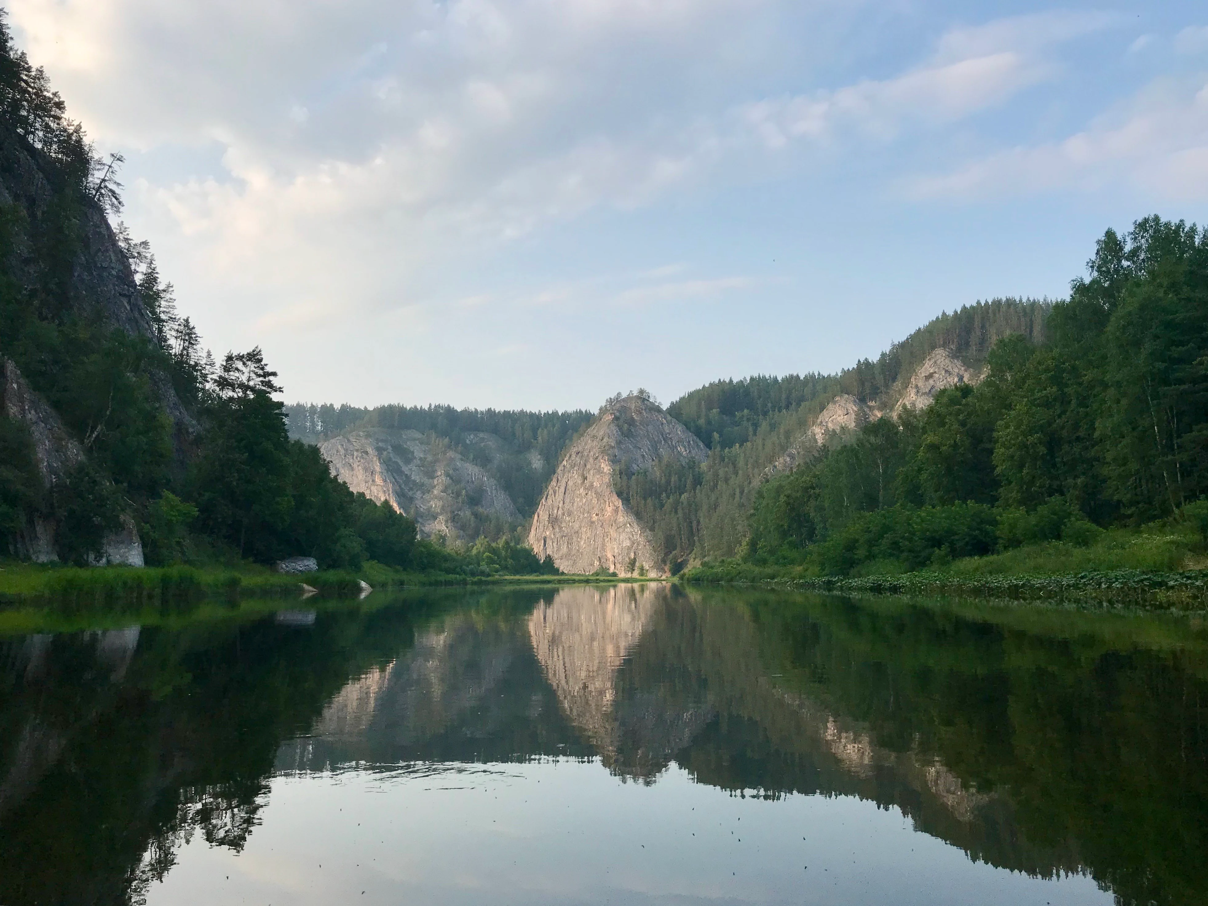 A wide river reflecting mountains and trees under a partly cloudy sky
