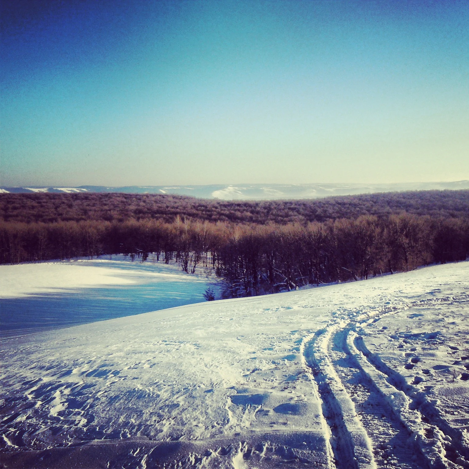 Snow-covered hills with tracks leading to a reservoir surrounded by forest under clear blue sky