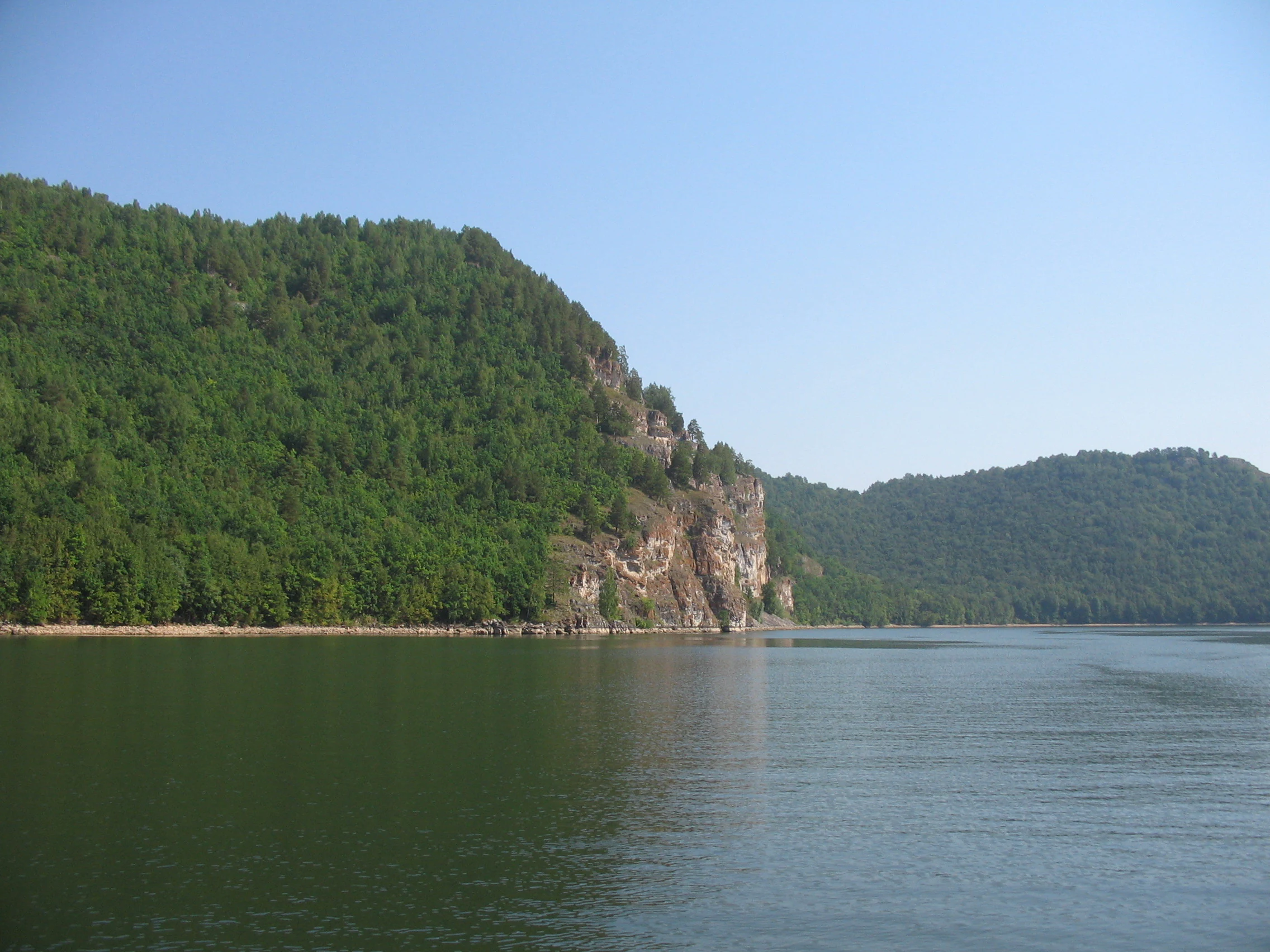 Calming water reservoir with lush green forested hills and rocky cliffs under a clear blue sky