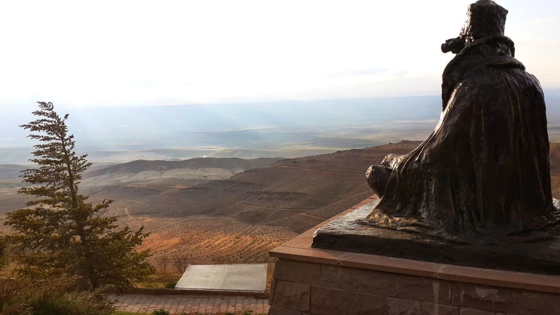 Bronze statue of a seated figure on a stone pedestal with a valley and hills in the background