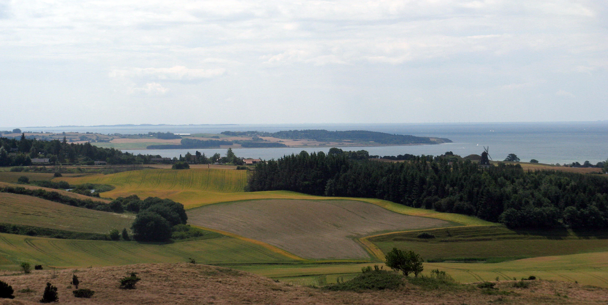A wide, elevated view of the Danish coast, featuring rolling fields, patches of forest, and the waters of Begtrup Vig under a cloudy sky.