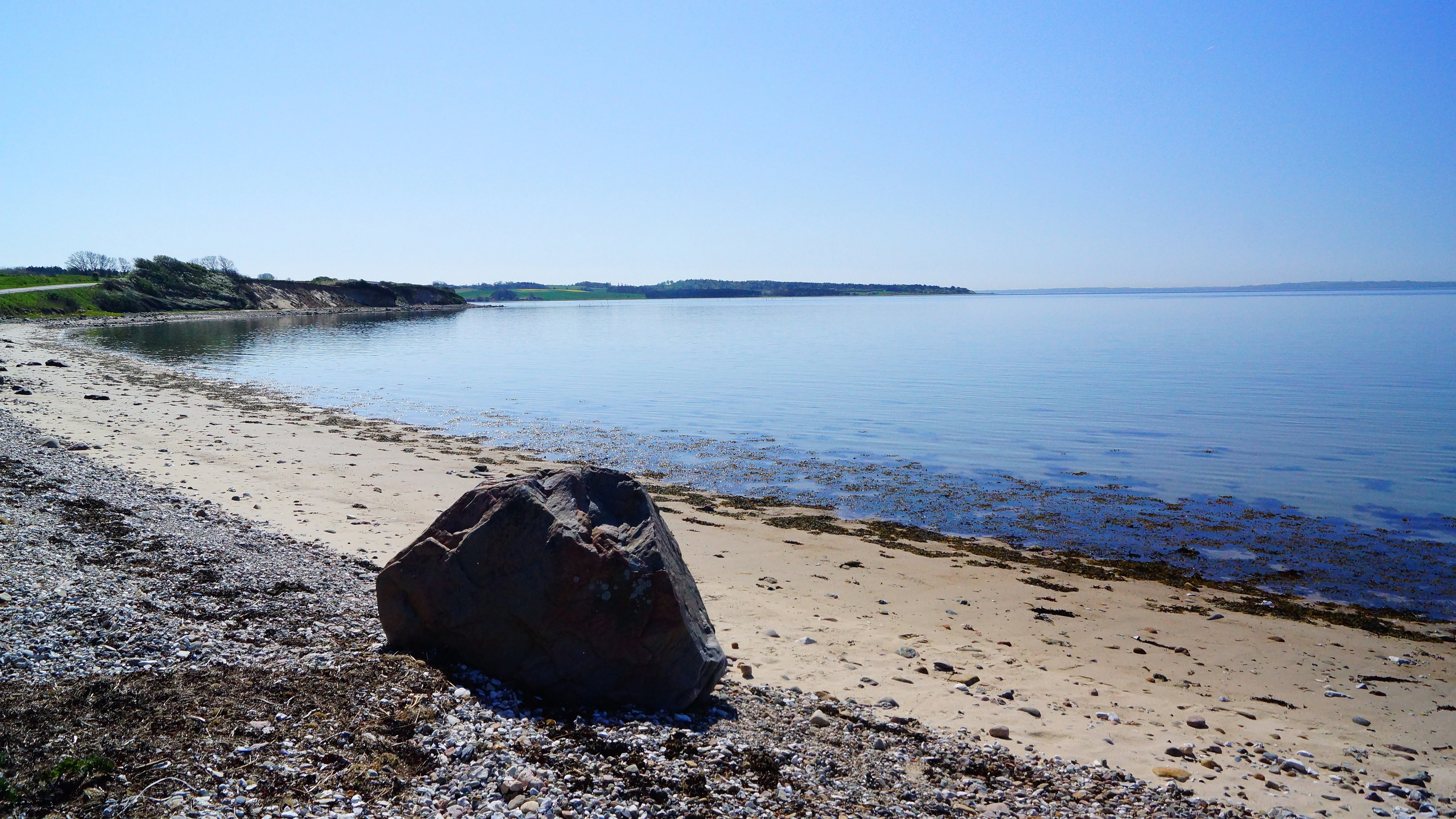 A wide coastal landscape showing a sandy beach and calm water under a clear blue sky, with a large boulder in the immediate foreground.