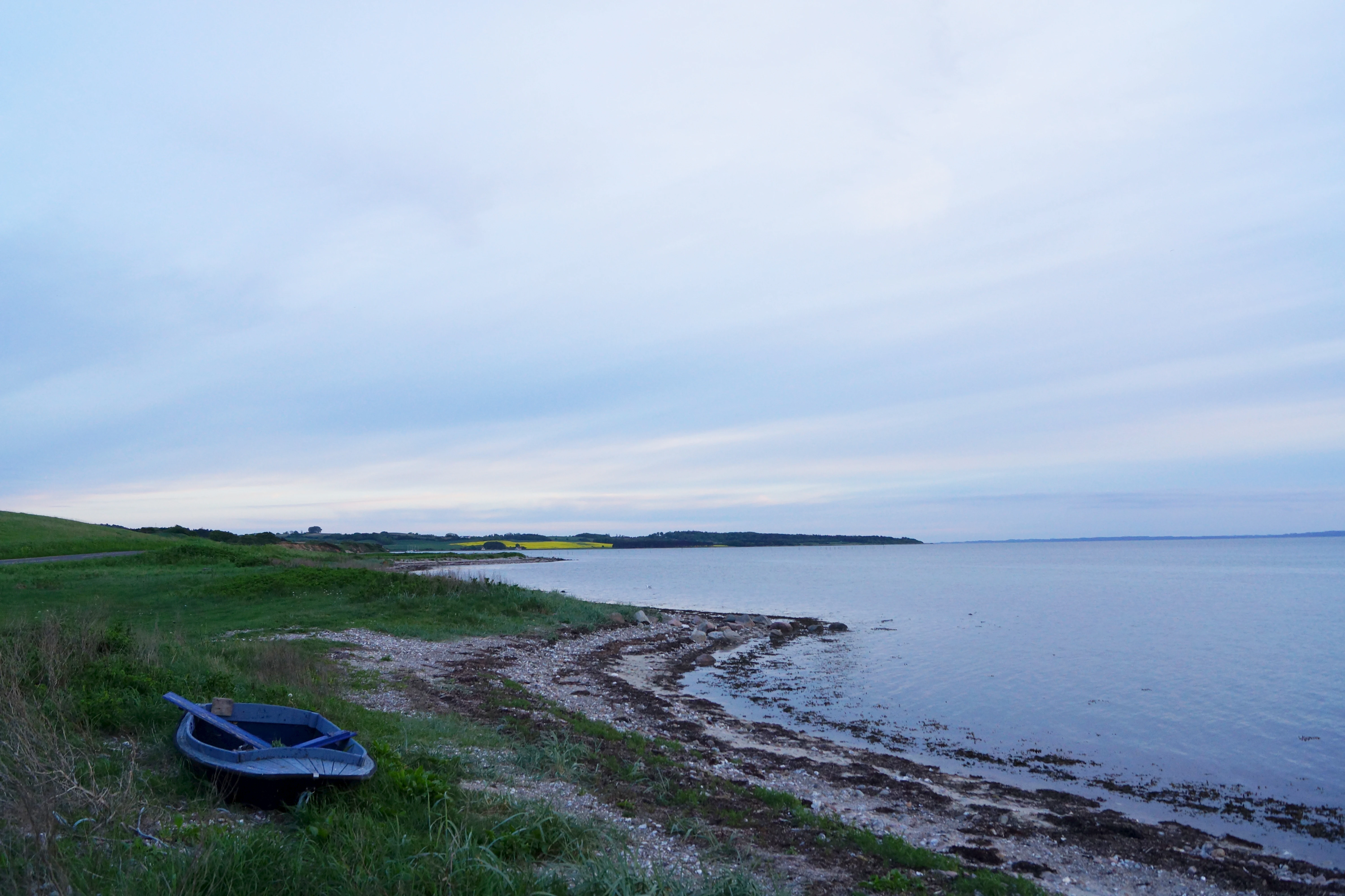 A small rowboat rests on the grassy shore of a calm, wide bay under an overcast sky.