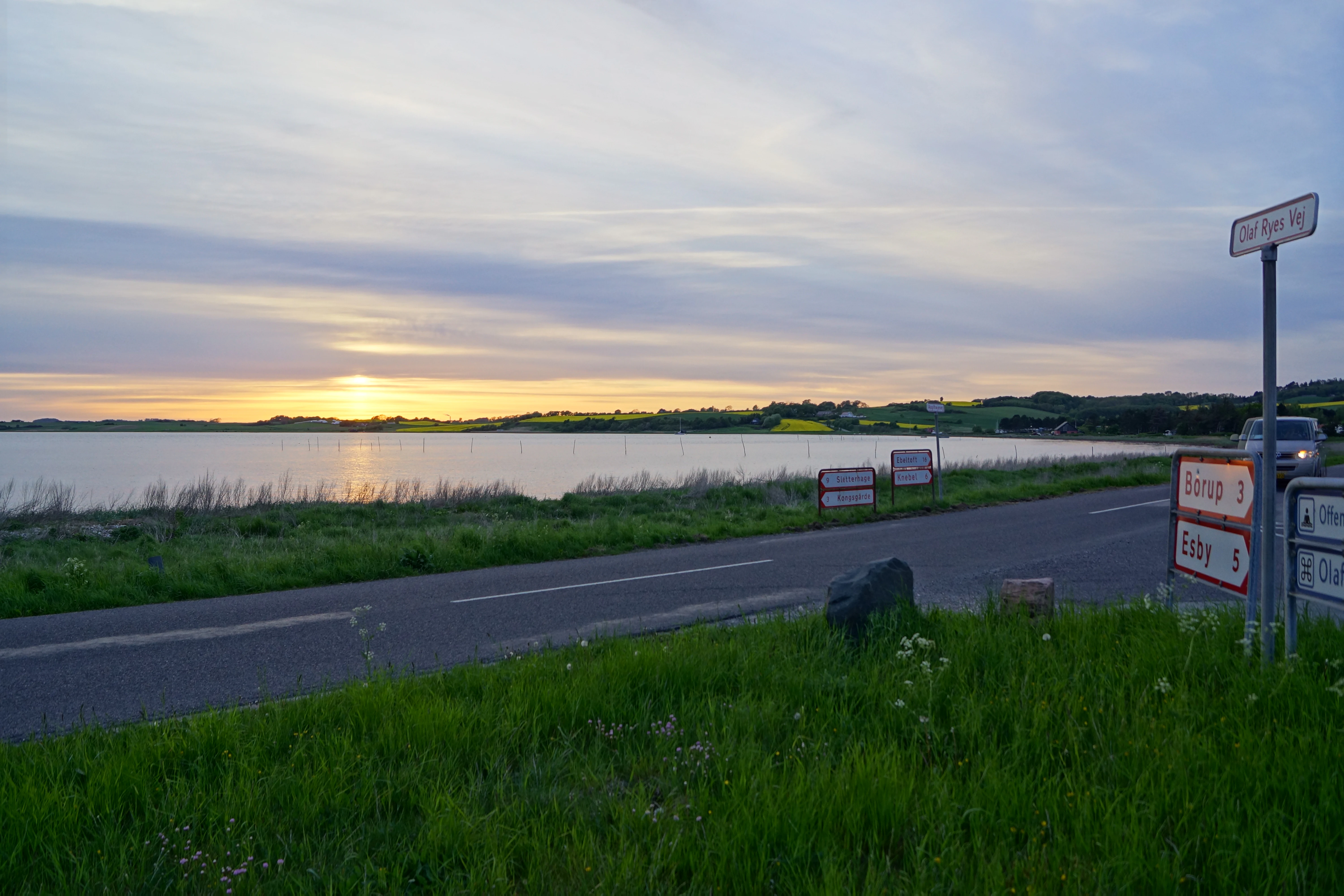 A wide shot showing a sunset over the calm waters of Begtrup Vig with a road running alongside the coast and several road signs on the right.