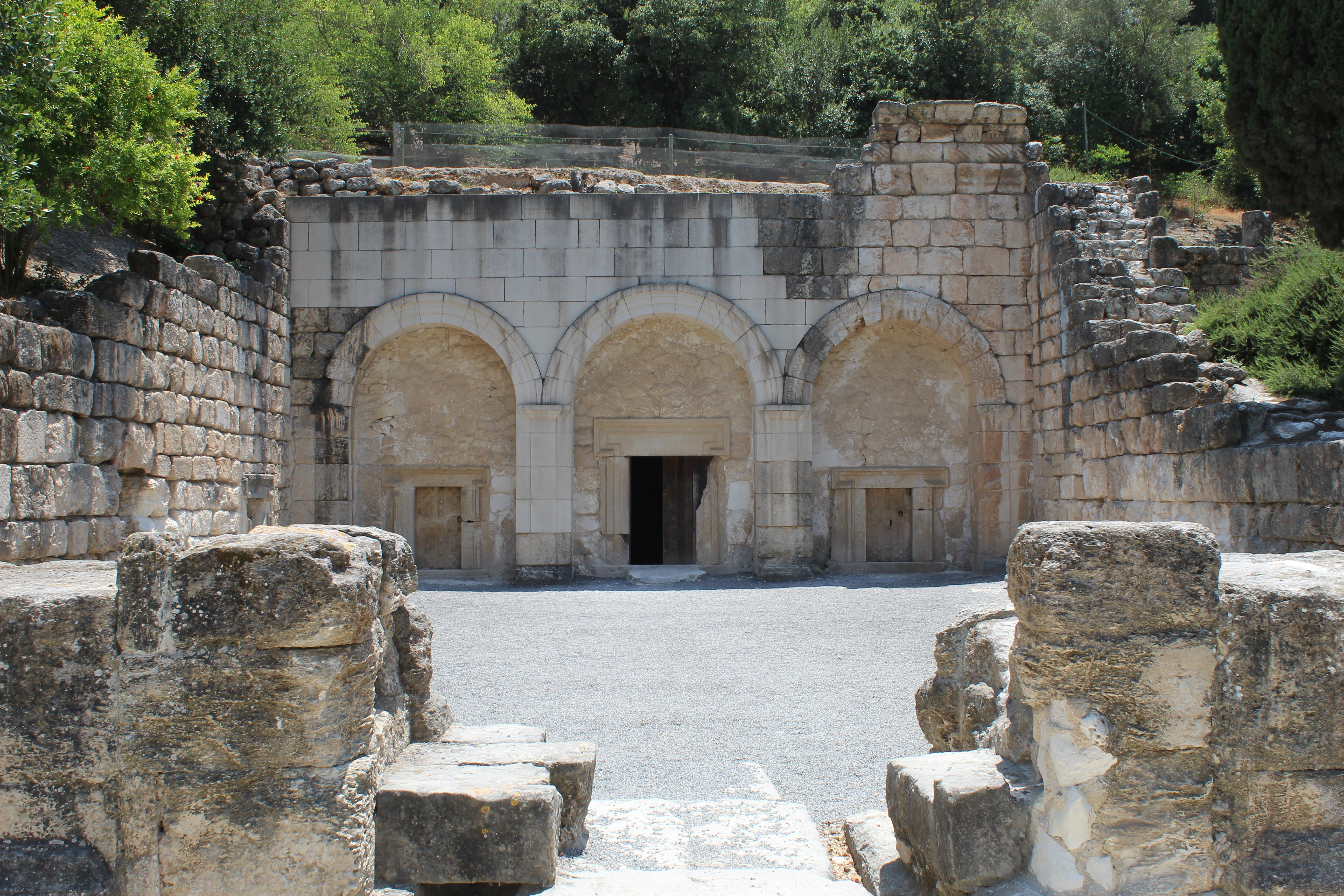 Stone arches and reconstructed walls of a burial cave entrance surrounded by ancient ruins and greenery