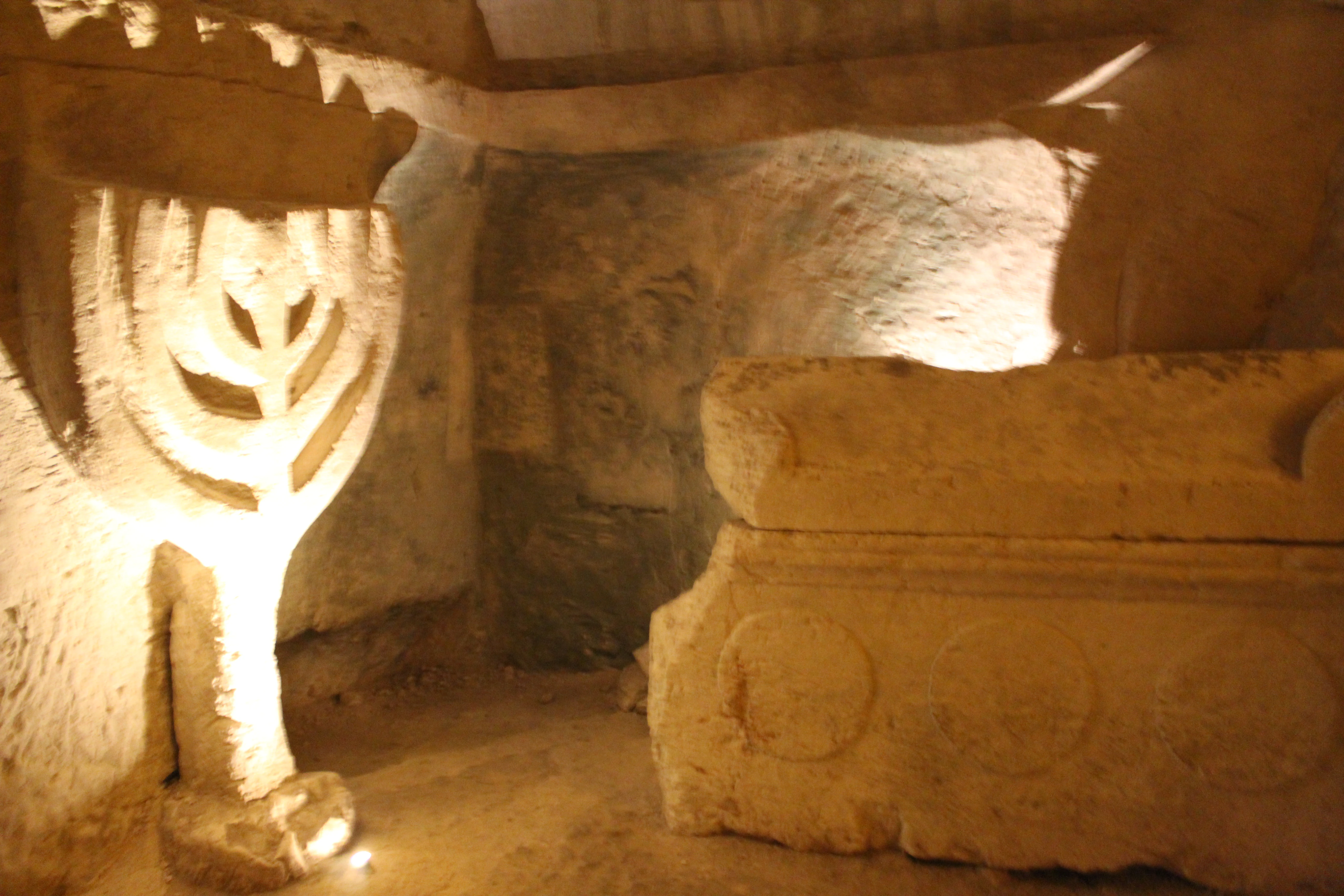 Stone menorah with seven branches and a sarcophagus in a carved cave chamber