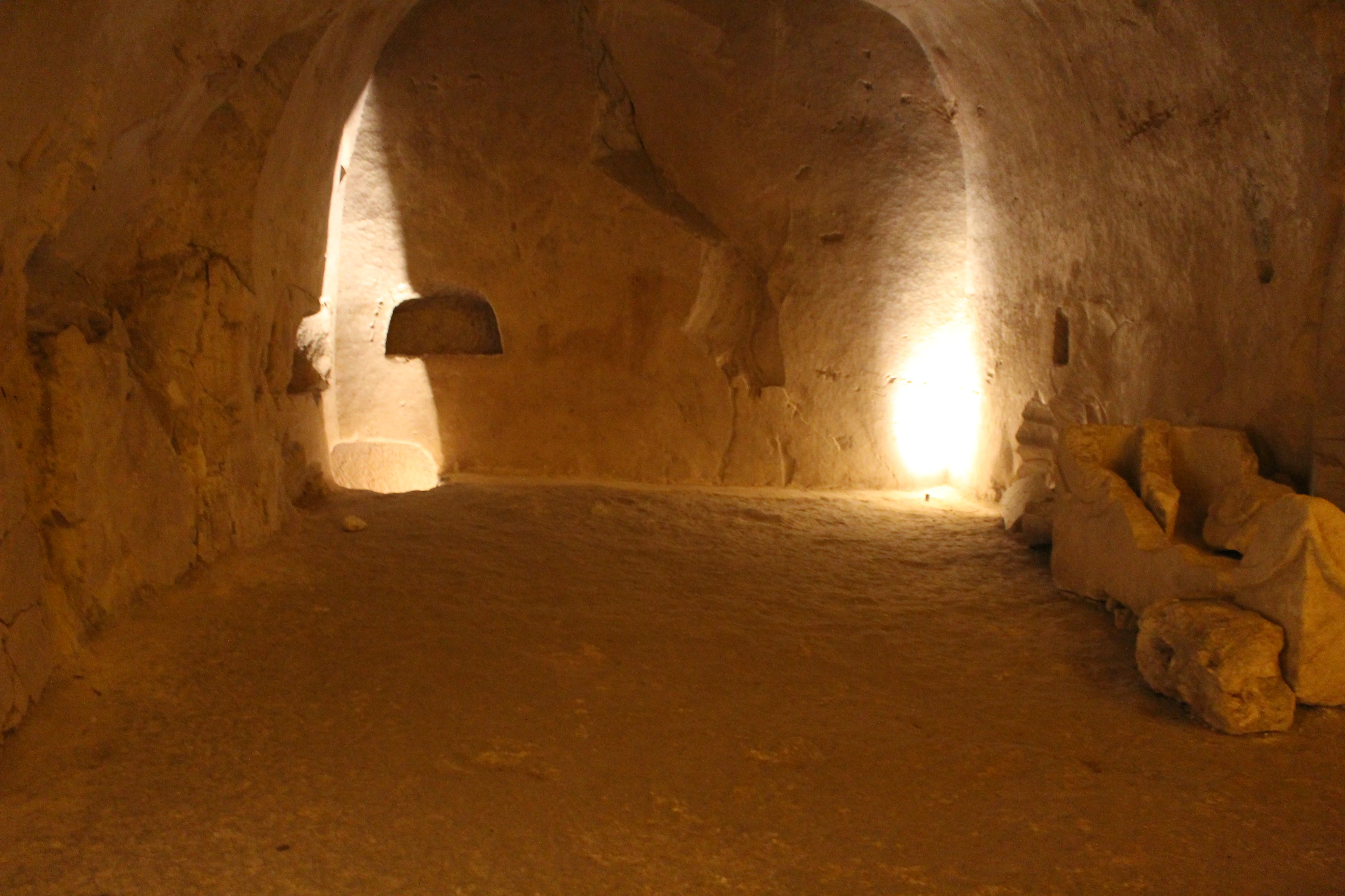 Stone-walled burial chamber with arched ceiling, illuminated by artificial light, showing a recessed niche and stone artifacts on the right