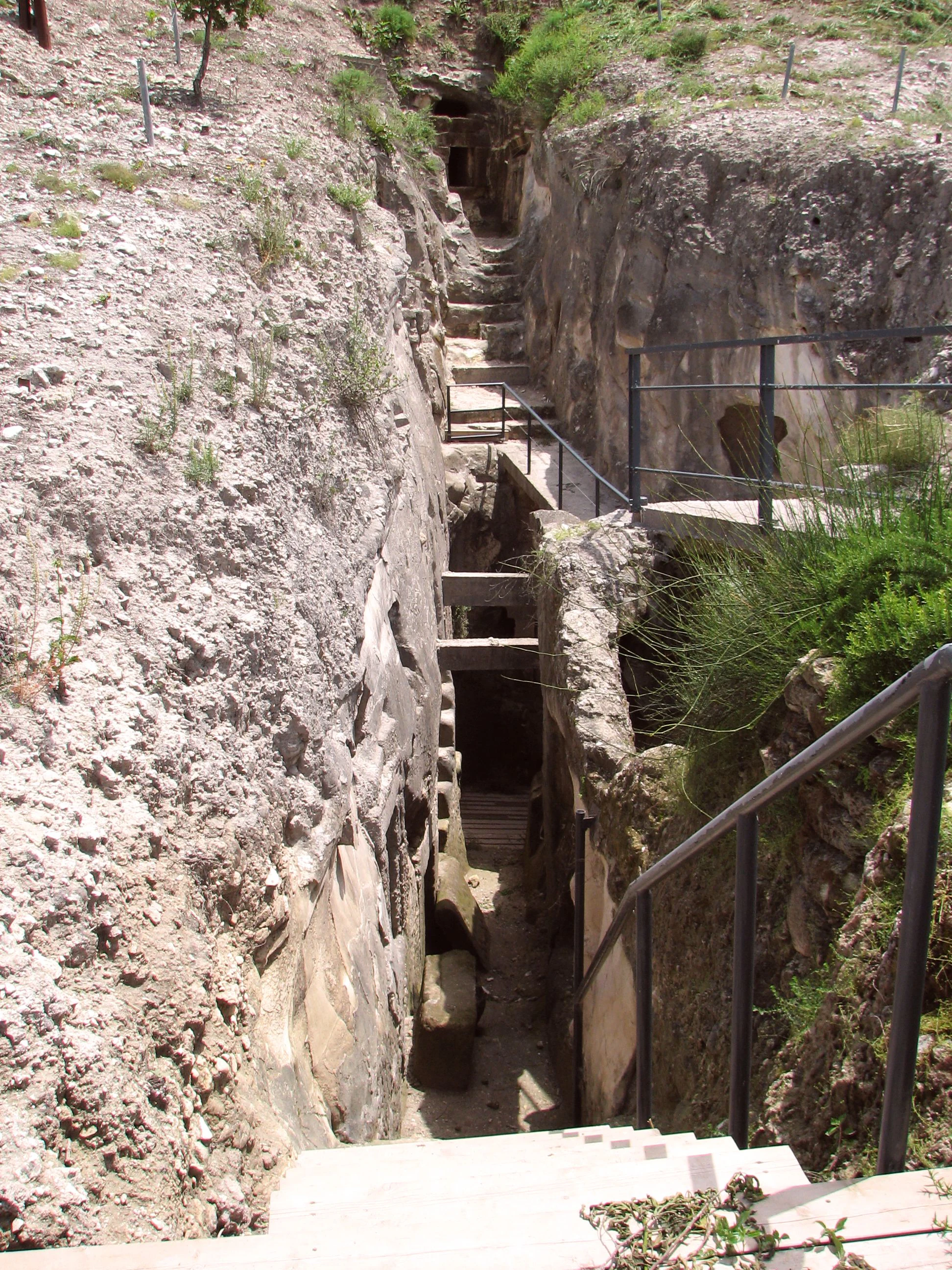 Stone steps and metal railings descending into a rocky trench with ancient cave entrances carved into the rock face