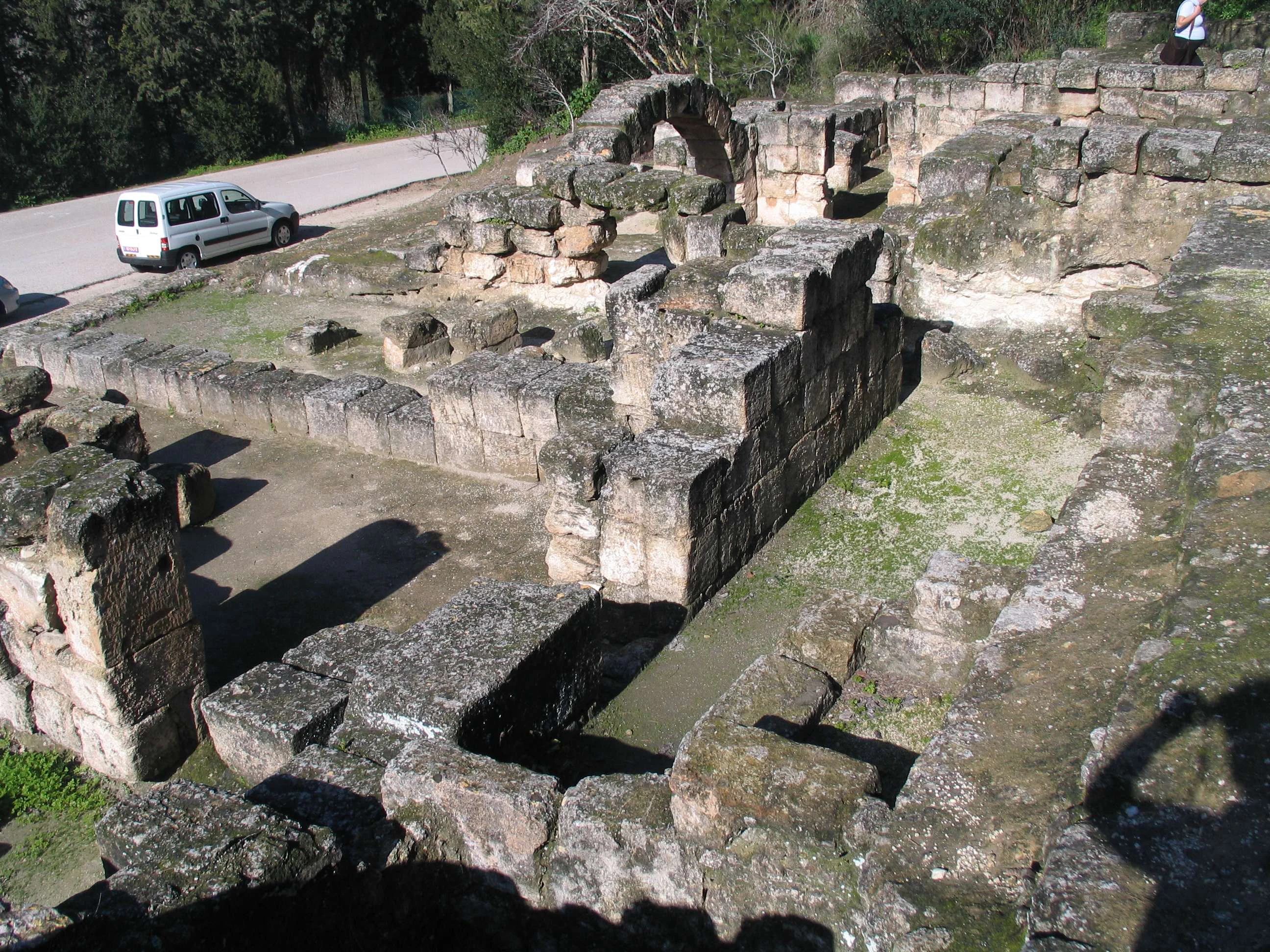 Stone ruins of the ancient synagogue with weathered walls and a paved area, surrounded by greenery and a road with a vehicle in the background.
