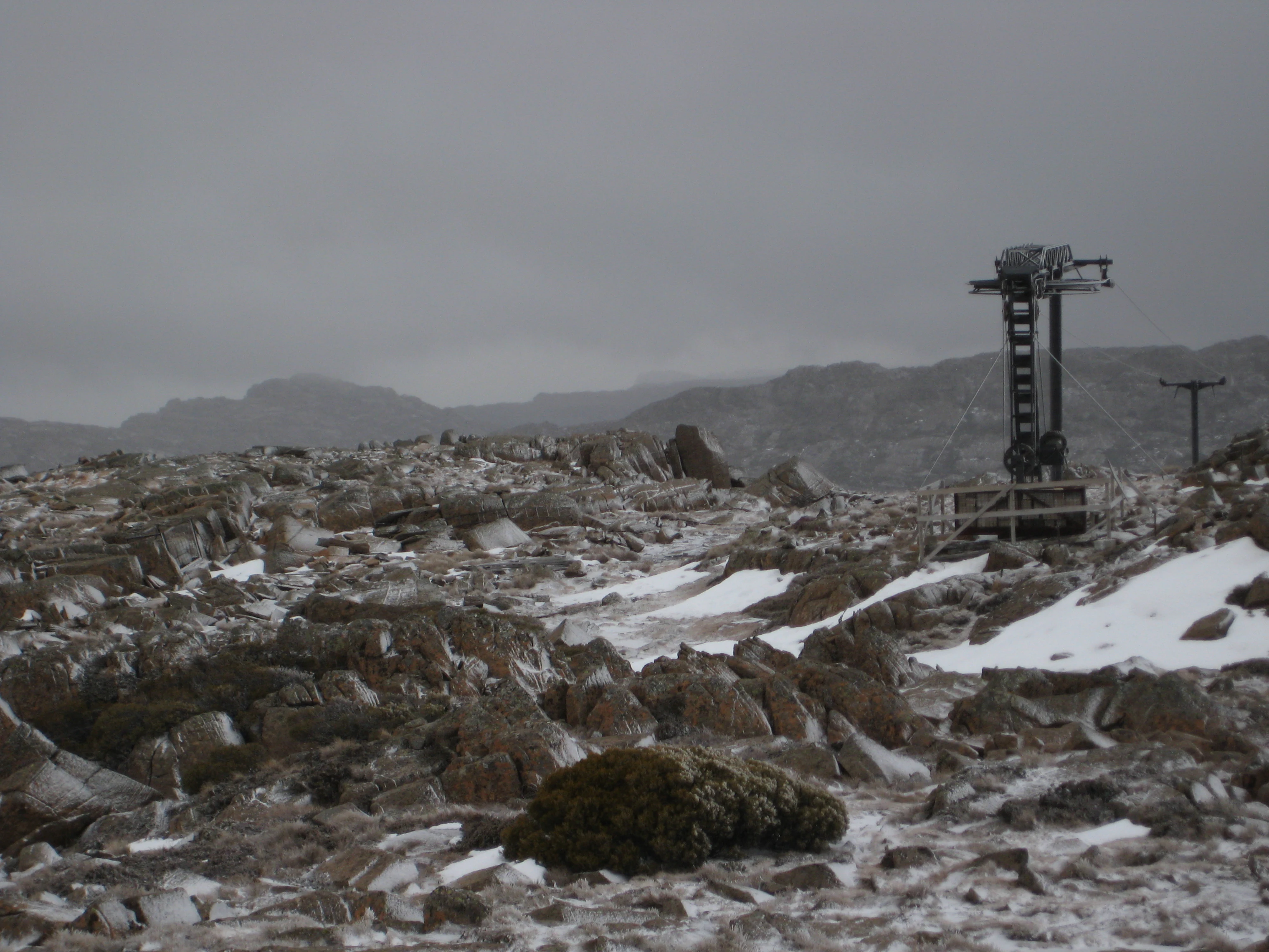 Terreno rocoso y nevado con rocas dispersas y vegetación escasa bajo un cielo nublado. Se aprecian una torre de telesilla y una valla a la derecha, con montañas lejanas al fondo.