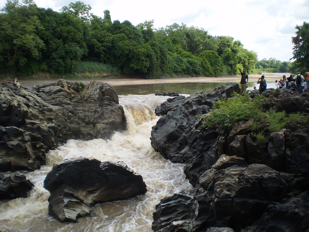 Rápidos fluyendo a través de rocas oscuras en el río Bénoué con exuberantes árboles verdes al fondo y un pequeño grupo de personas observando desde la orilla del río