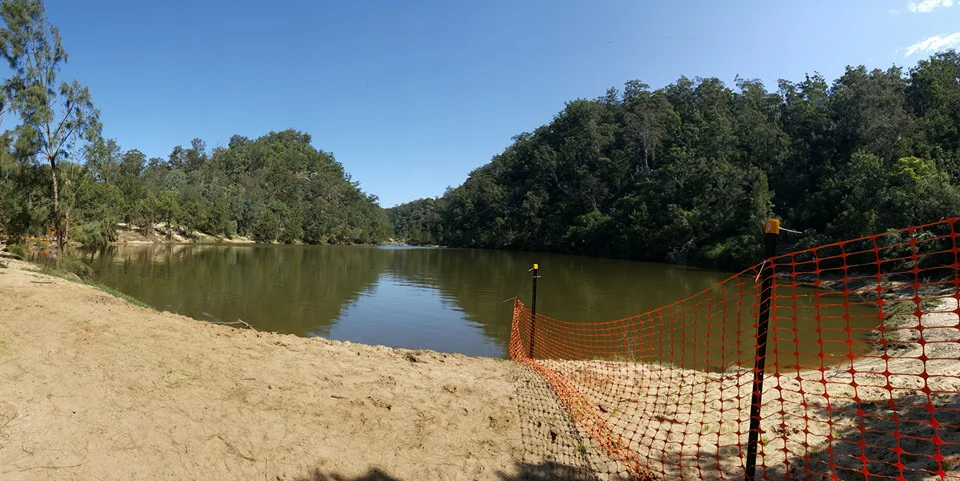 A wide view of a calm river basin with a sandy beach in the foreground and a orange safety fence bordering the water.