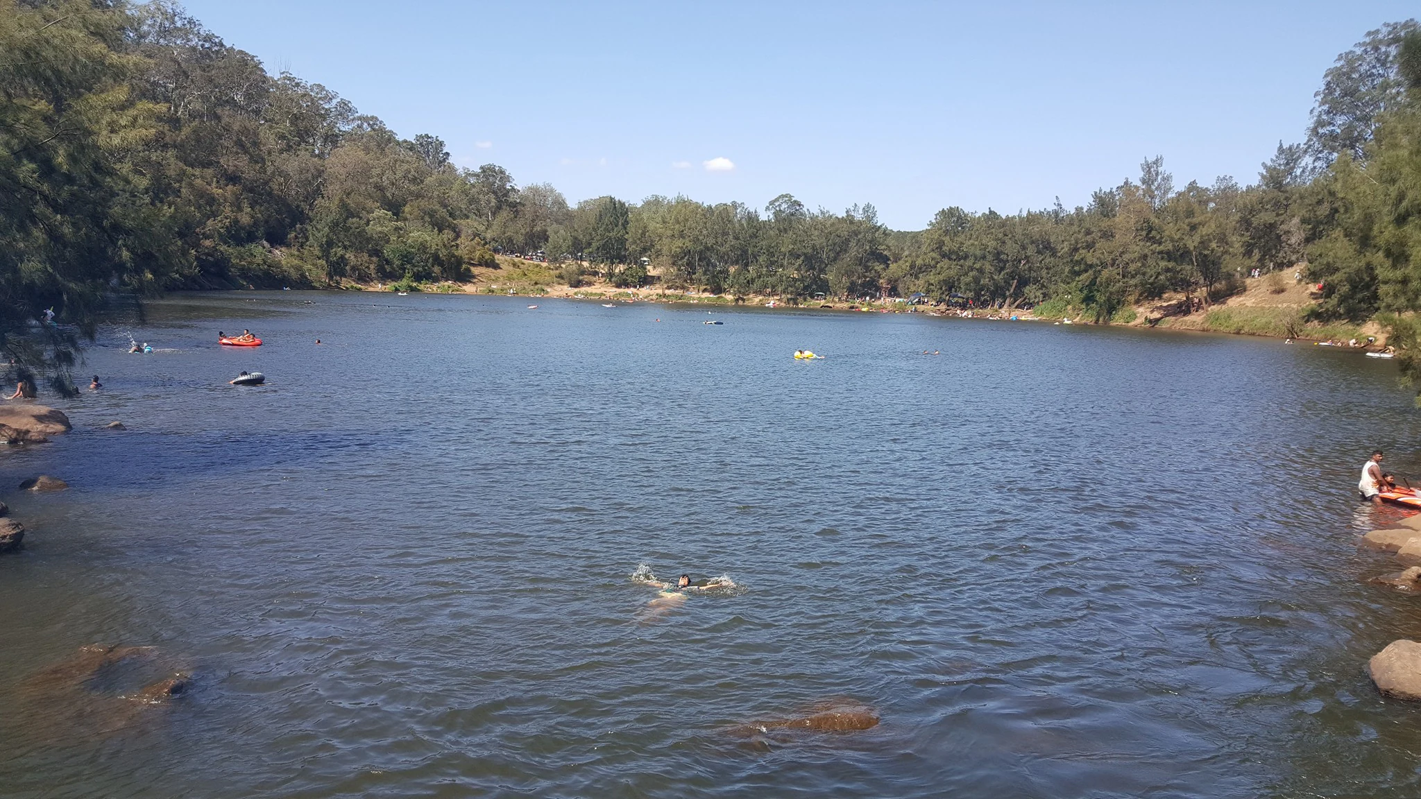 A wide view of a large natural water basin surrounded by dense trees and rocky shores, with several people swimming and using small inflatables in the water.