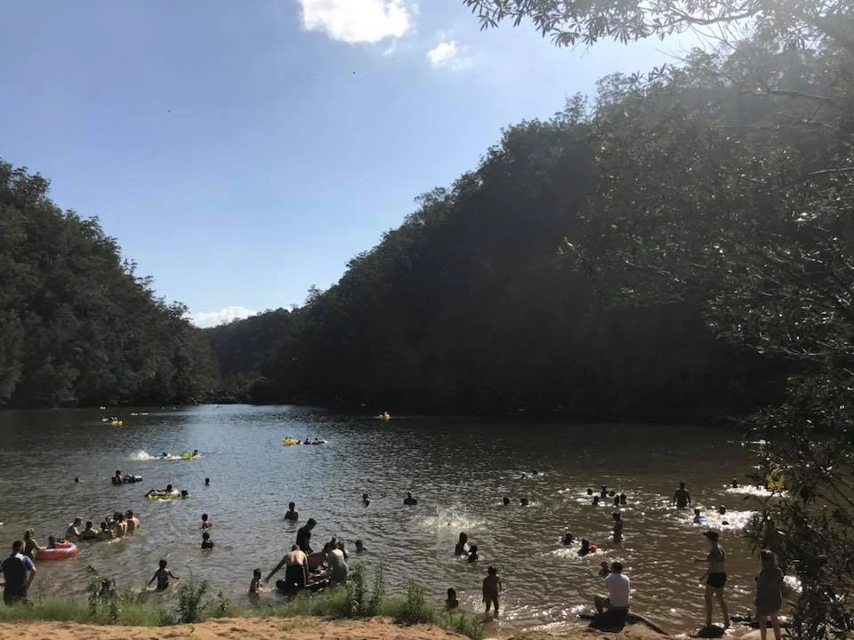 A wide shot of a swimming hole surrounded by forest, with many people swimming and playing in the water on a sunny day.