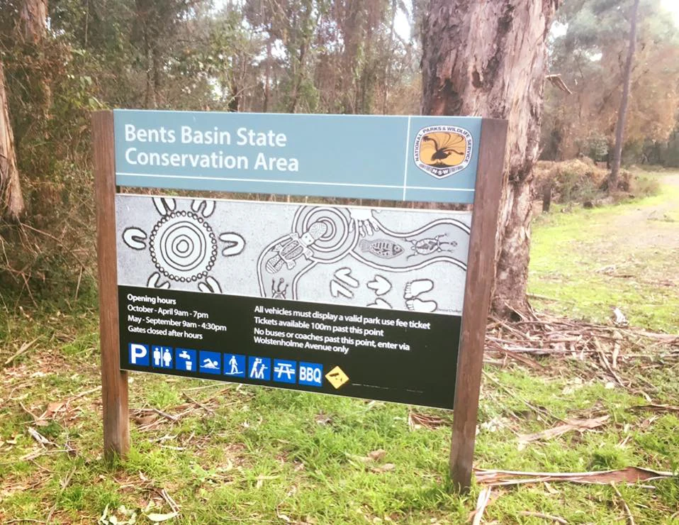 A metal park entrance sign for Bents Basin State Conservation Area standing in a wooded area, featuring informational text, icons for park facilities, and Indigenous-style artwork.