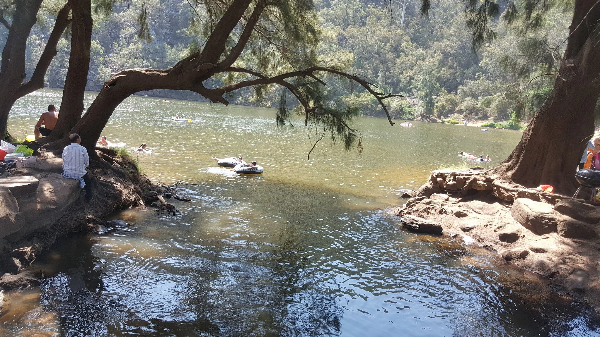 A group of people relaxing and swimming in a wide river section, with large trees overhanging the riverbank.
