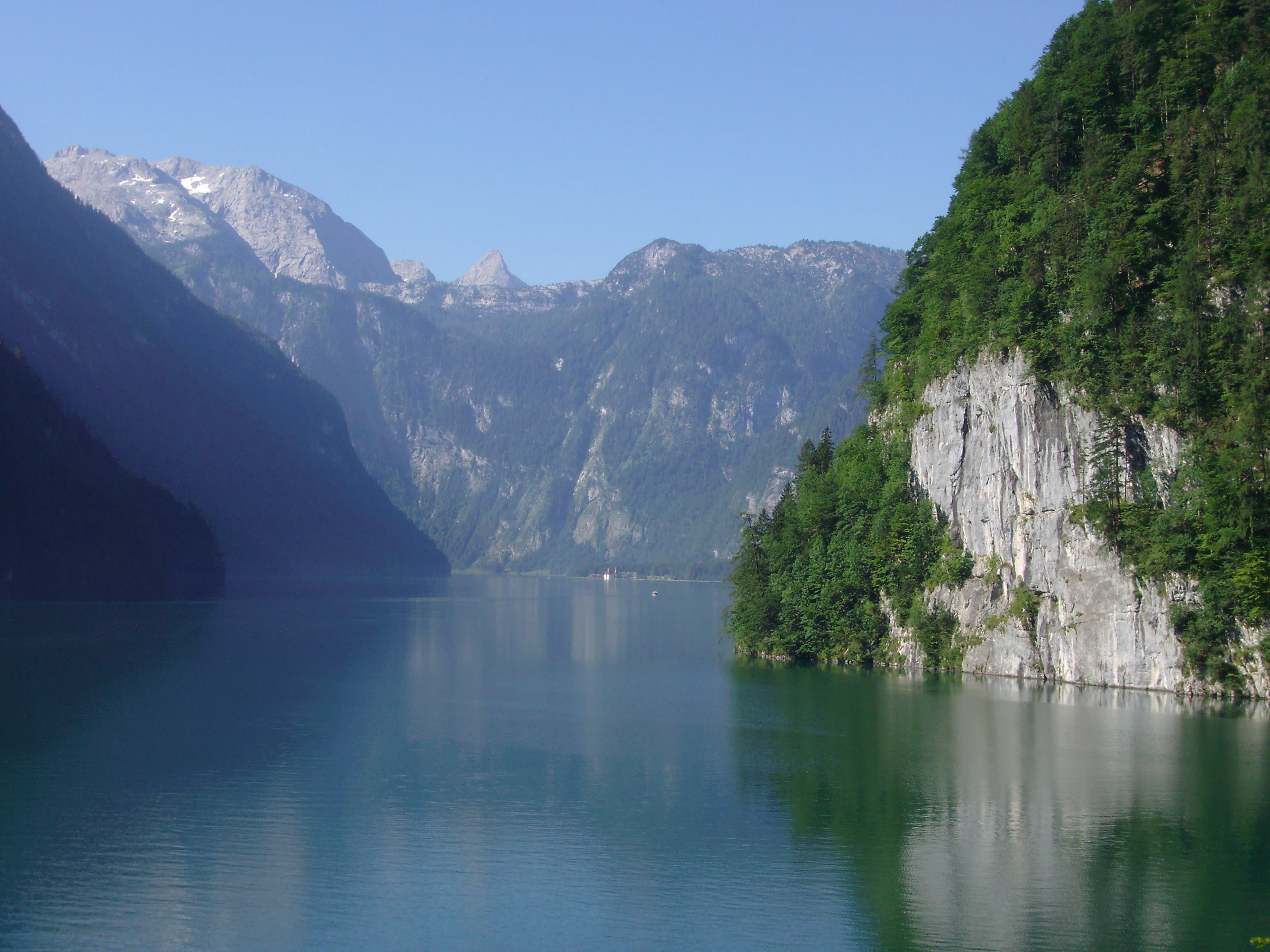 Calm turquoise lake surrounded by steep rocky cliffs and lush green mountains under clear blue sky