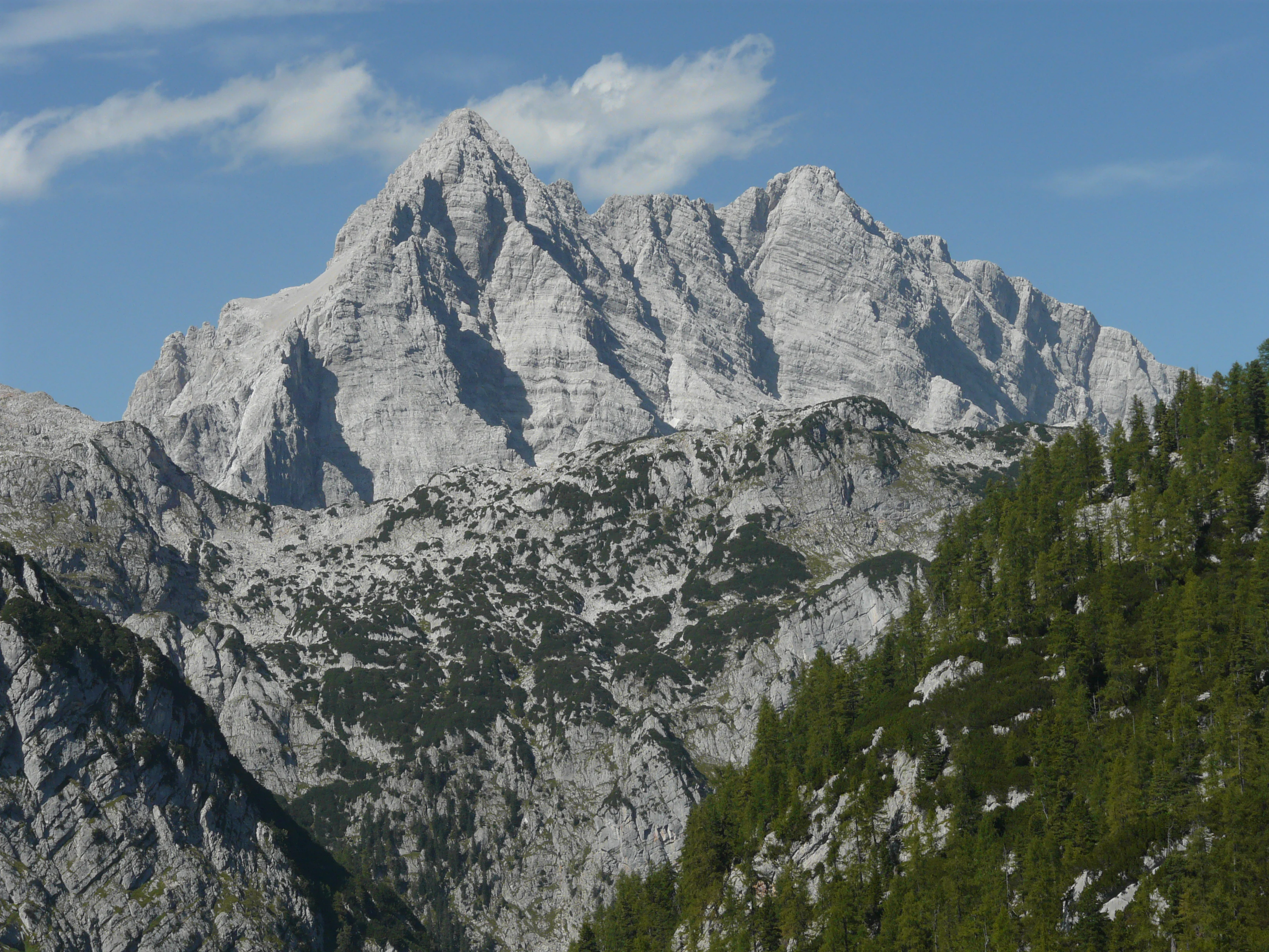Large rocky mountain peak with steep slopes and forested hills under a clear blue sky
