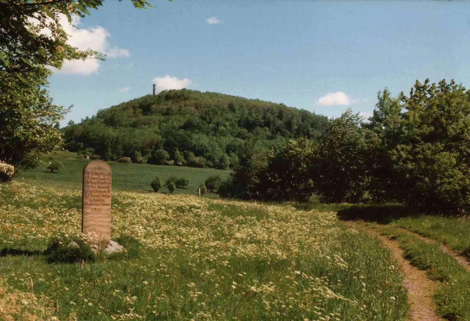 Un marcador de piedra se alza en un prado cubierto de hierba con un sendero de tierra, mientras que una colina boscosa con una torre en la cima surge al fondo bajo un cielo azul.