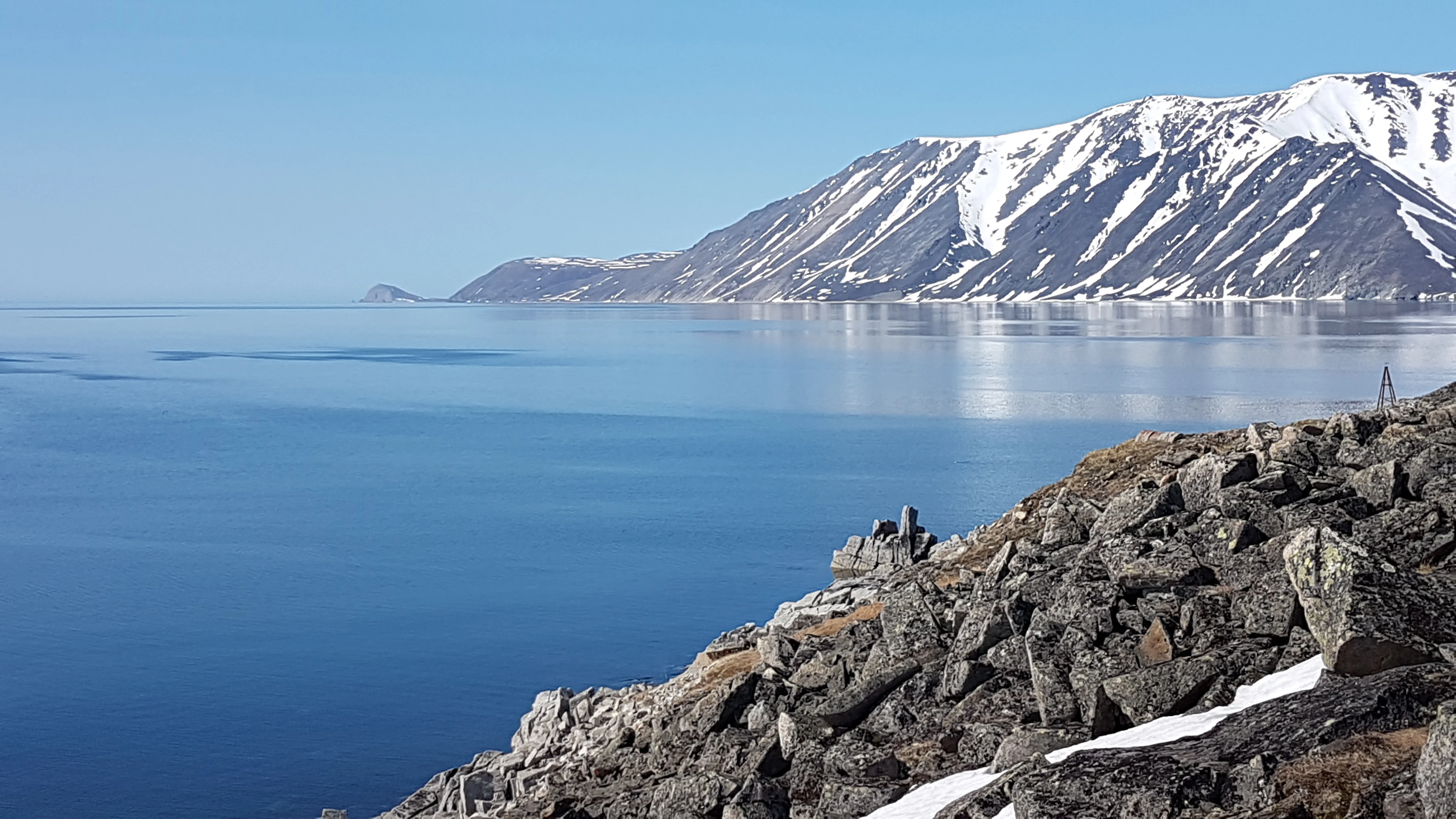 Rocky shoreline with patches of snow overlooking a calm blue bay, snow-covered mountains in the distance under a clear sky
