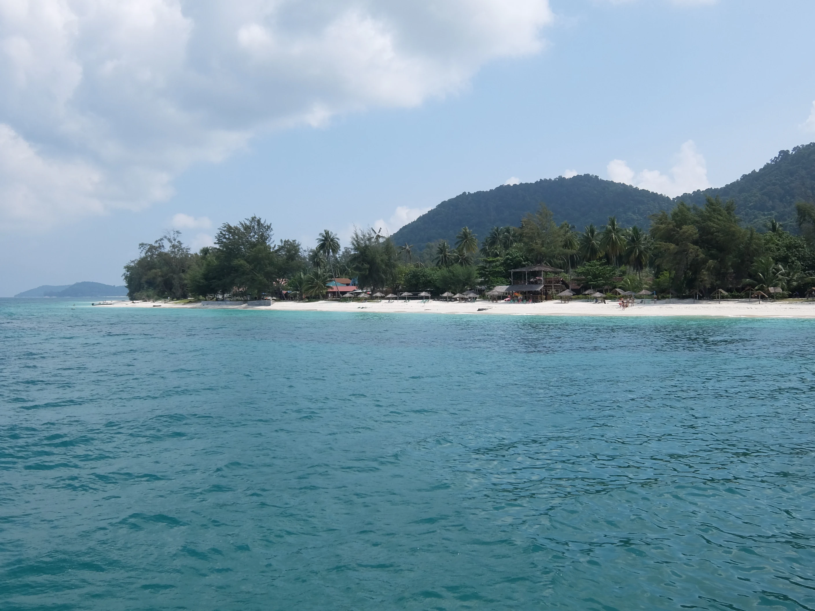 A wide angle view from the water looking toward a white sand beach backed by dense tropical trees and a forested mountain on Besar Island.