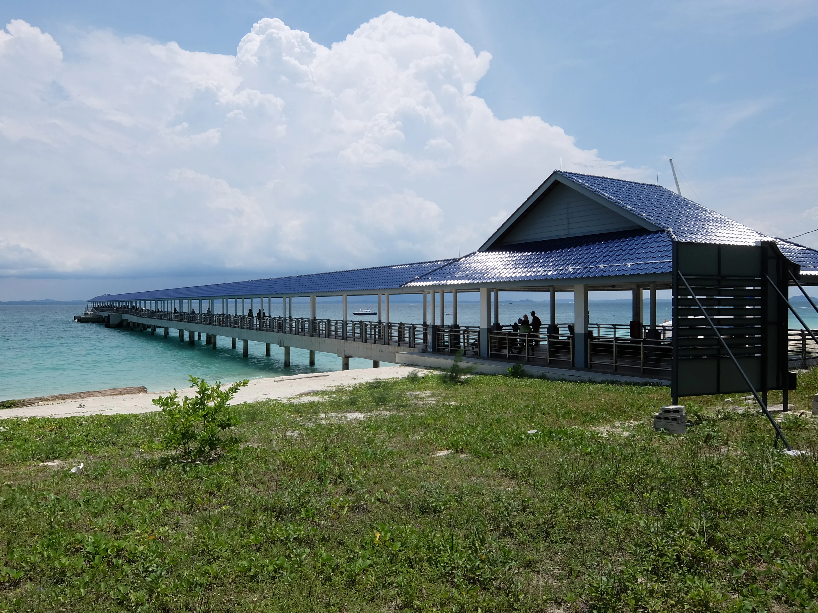 A long, covered wooden jetty with a blue tiled roof extending from a grassy beach area into the turquoise ocean under a cloudy sky.
