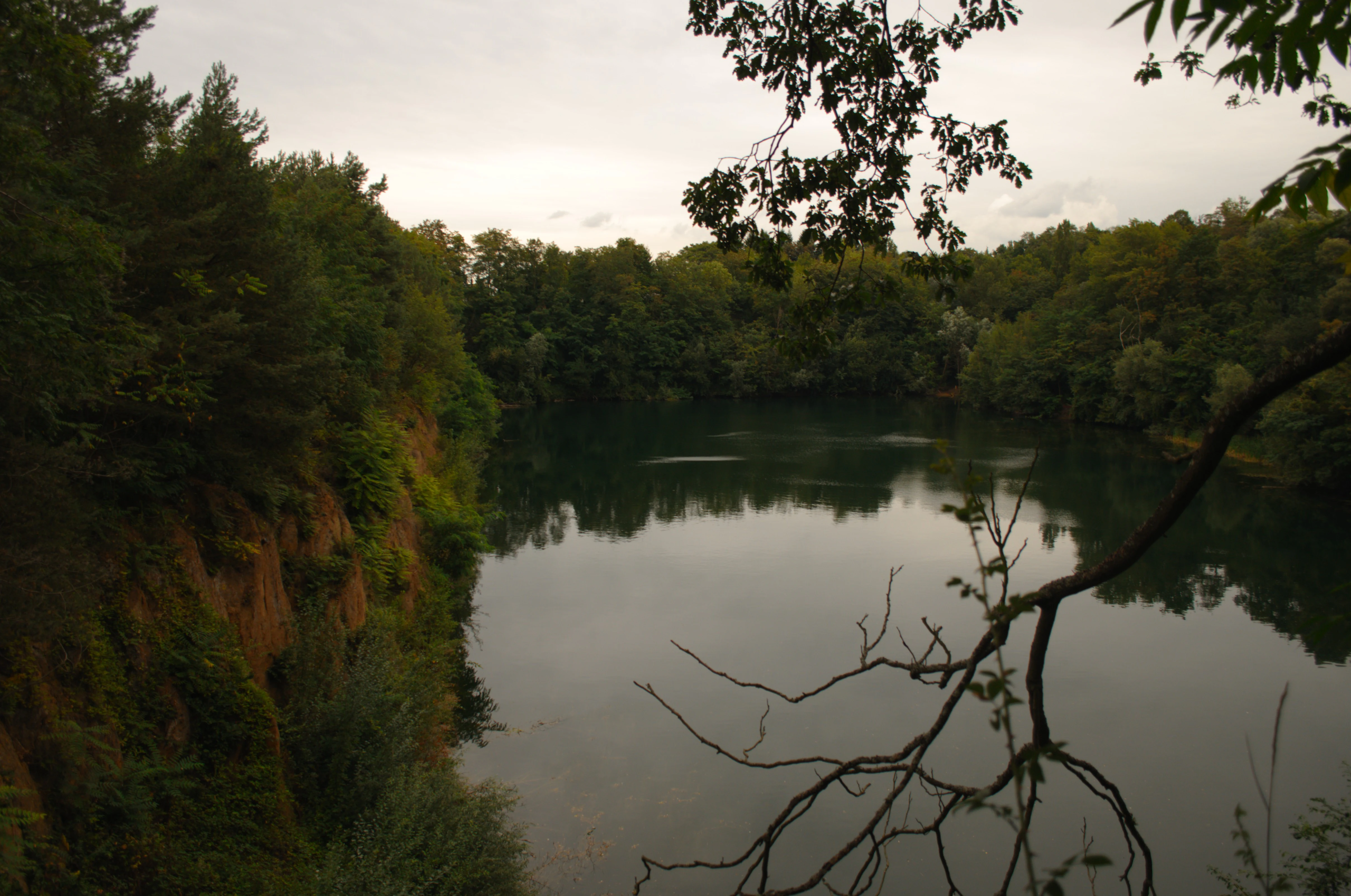 A calm, dark lake situated in a deep basin with steep earthen embankments and dense surrounding forest, viewed from a slightly elevated position.