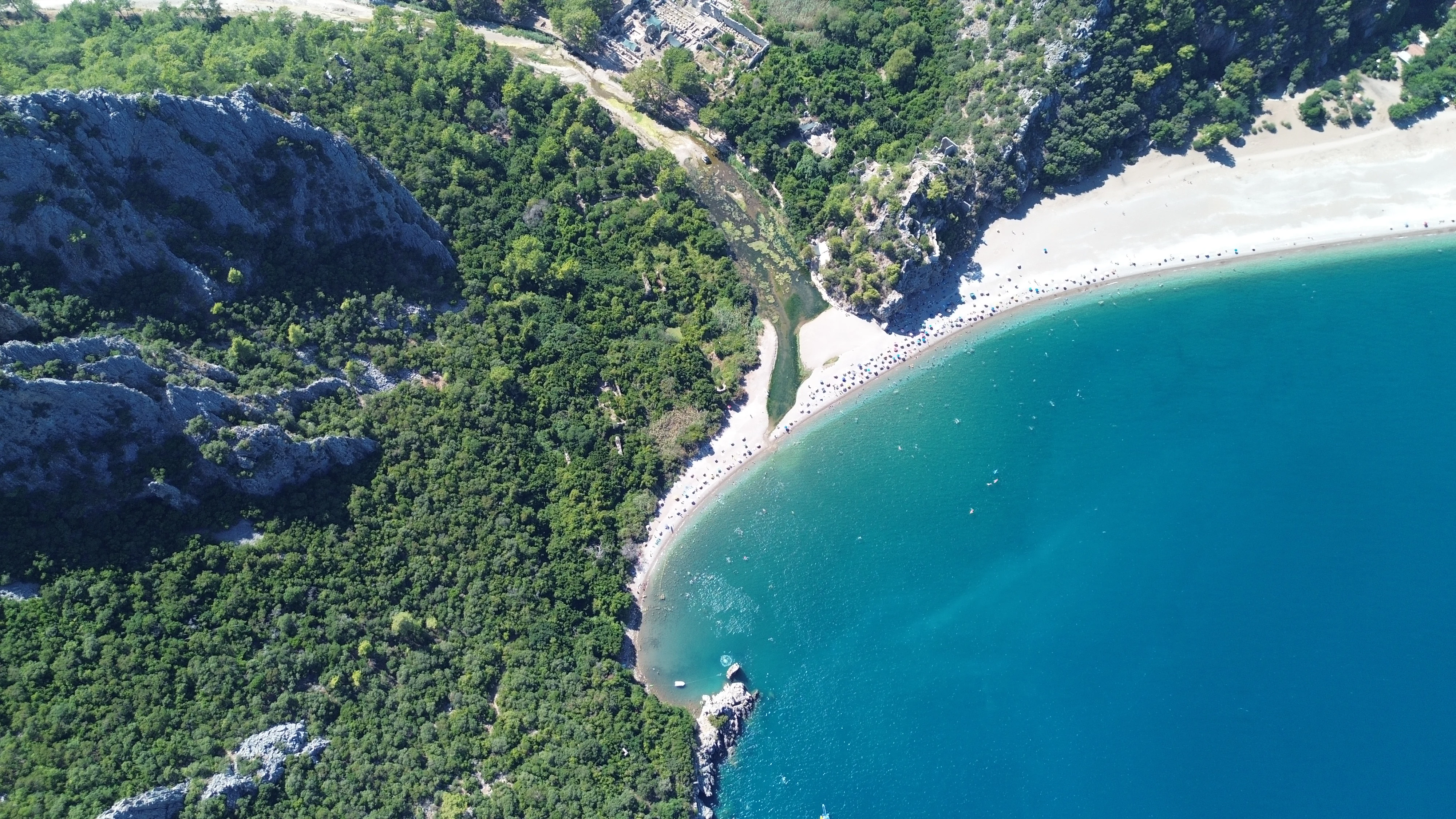 Aerial photograph of a coastal landscape featuring a sandy beach, clear blue water, rocky cliffs, and dense green forest along the shoreline.