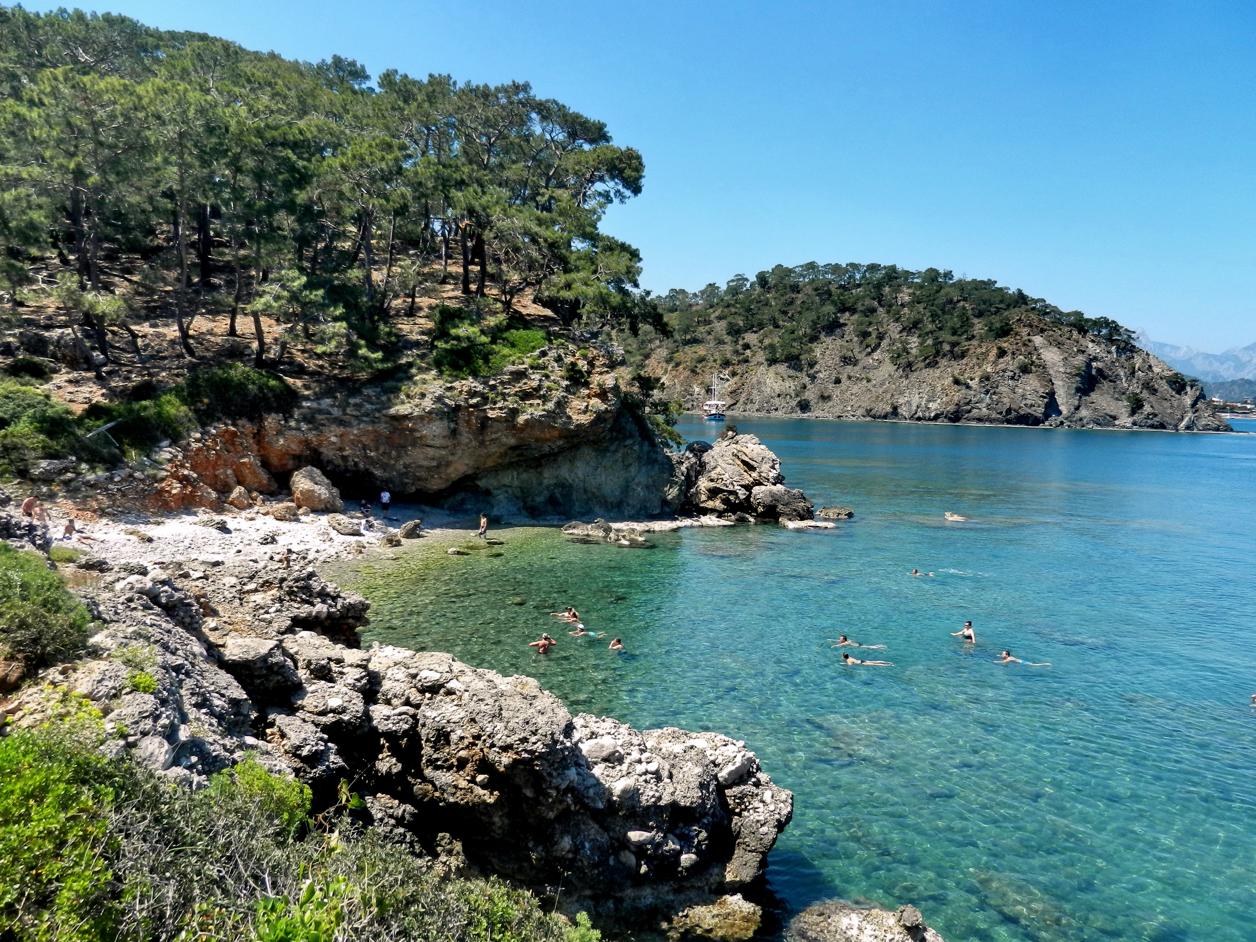 Rocky coastal bay with clear blue water, people swimming, forested cliffs, and a clear blue sky