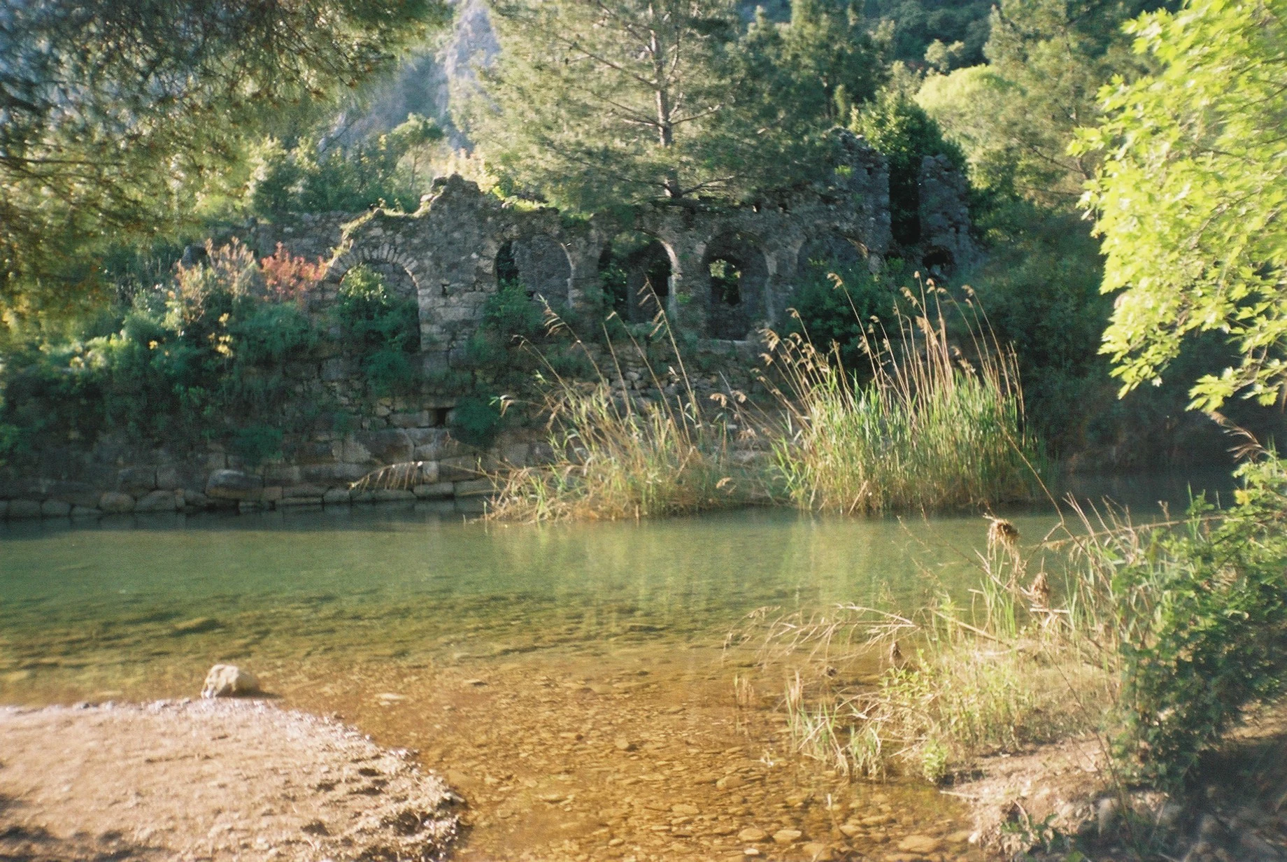 Ancient Roman bath ruins near a calm body of water with tall reeds and trees in the background