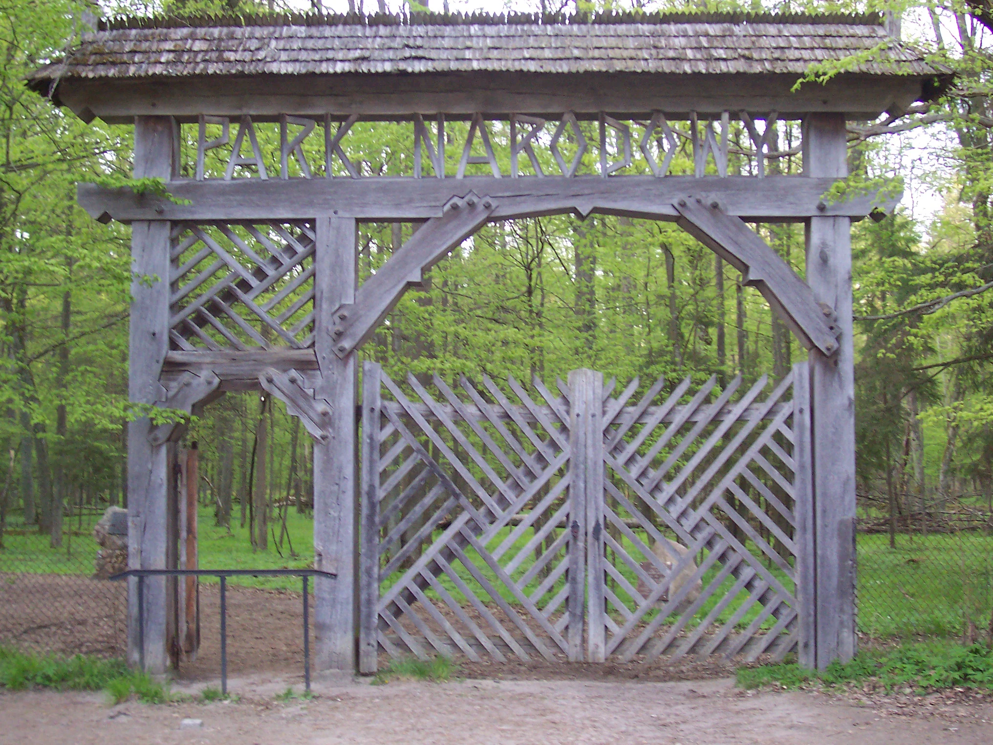 A large, traditional wooden gate with a shingled roof and lattice details, standing at the entrance to a forest in Białowieża National Park, with the words PARK NARODOWY carved into the top crossbeam.