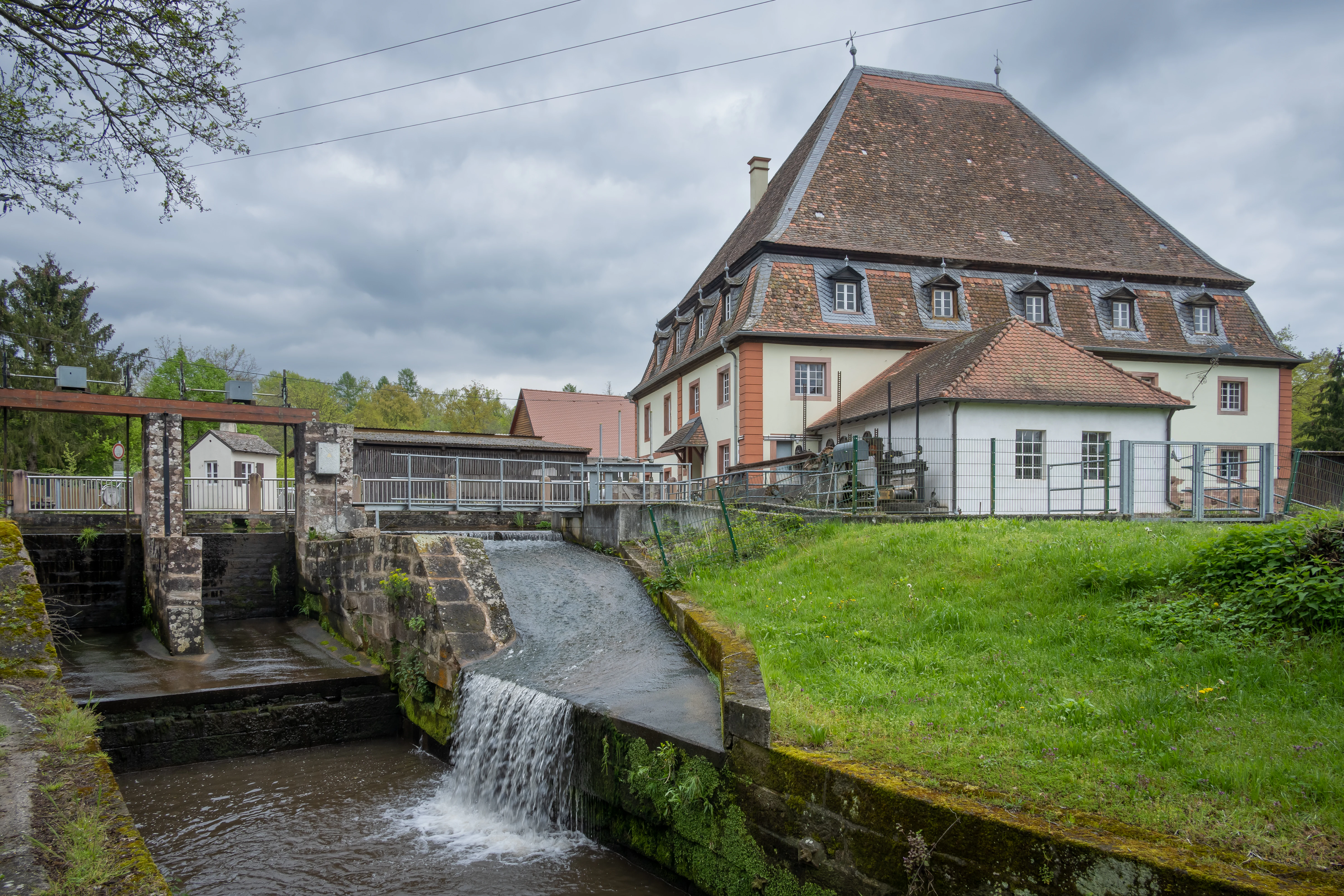 A multi-story historic mill building with a steep tile roof, featuring a small waterfall flowing into a stone-lined channel in the foreground.