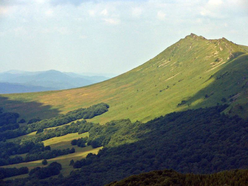 A sunny mountain peak with grassy slopes and forested lower hills under a partly cloudy sky.