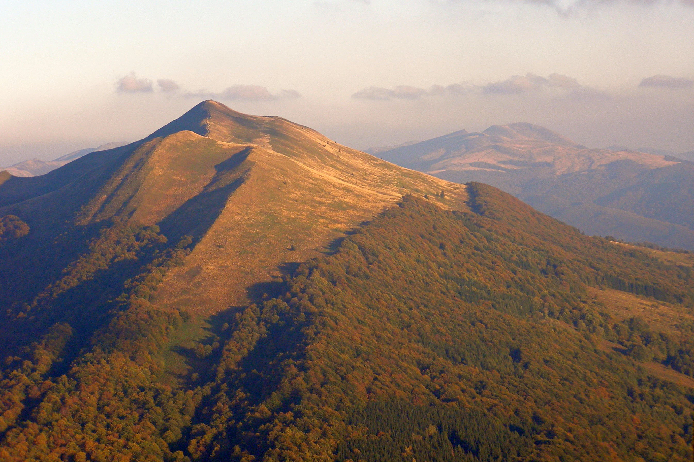 A wide aerial view of the Polonina Carynska mountain ridge, showing golden-brown grassy slopes above dense autumn-colored forests under a clear sky.