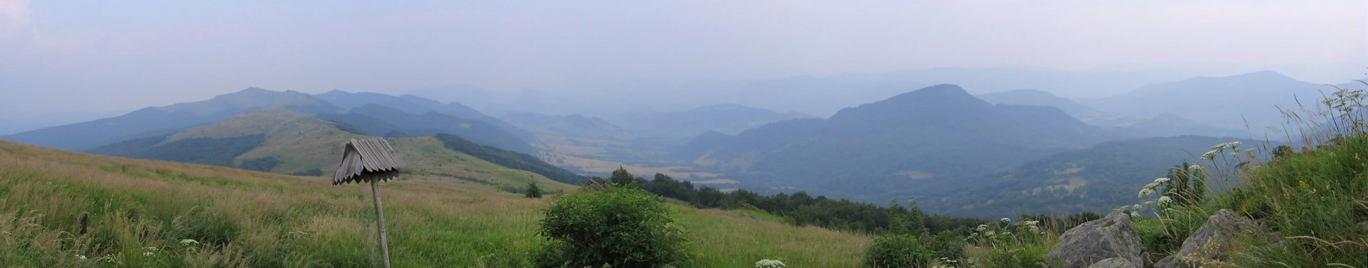 A wide panoramic landscape view of rolling, grassy mountain ridges under a hazy, overcast sky.