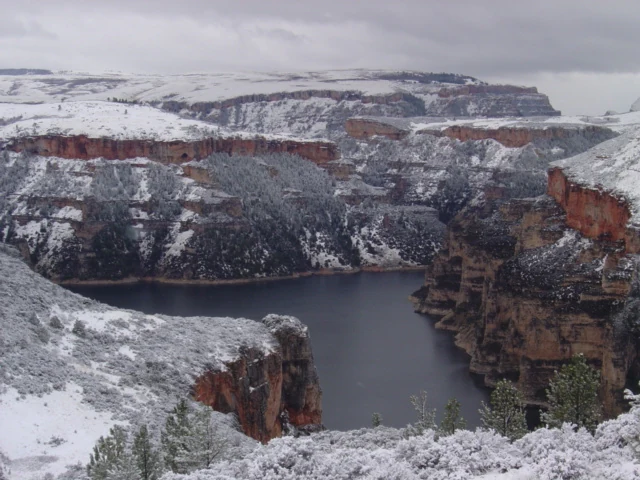 A wide scenic shot of a deep canyon with reddish-brown rock cliffs partially covered in snow under a grey, overcast sky. A dark, calm body of water occupies the canyon floor.