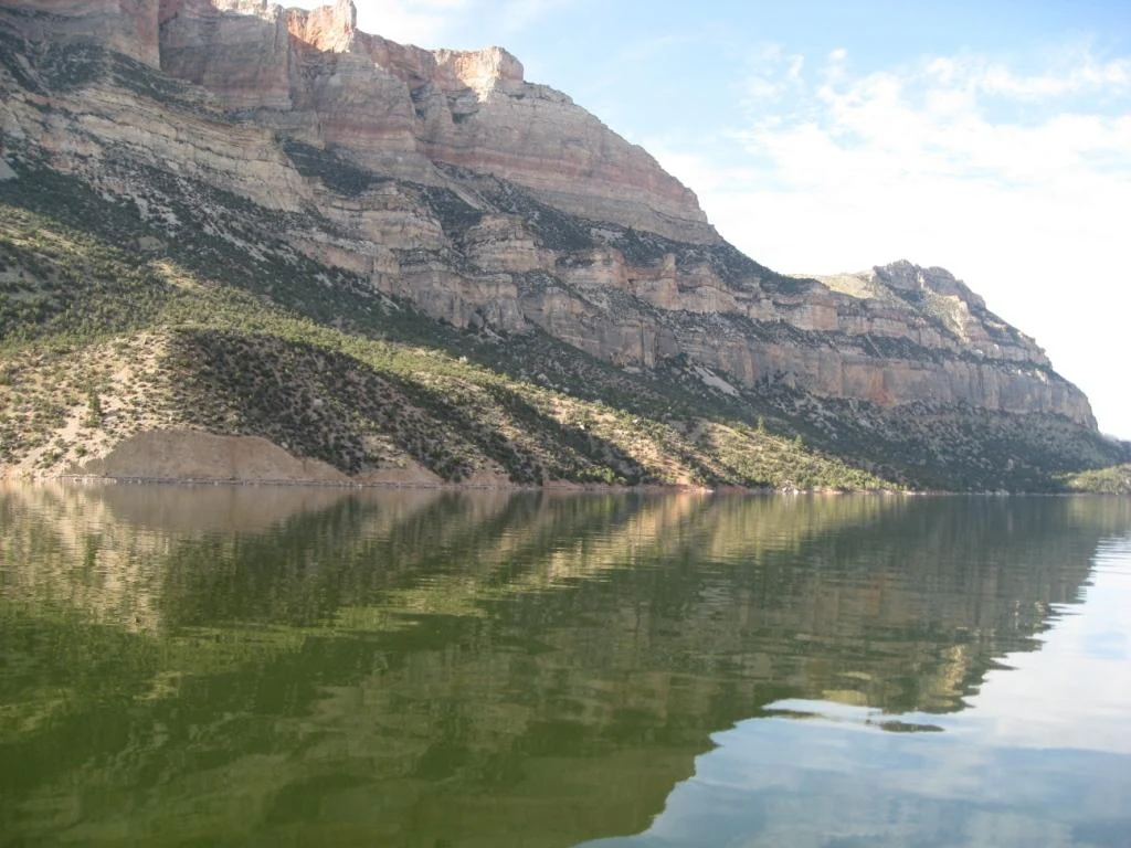 A wide, eye-level view of Bighorn Lake with massive, layered canyon cliffs towering over the water, creating a mirror-like reflection on the surface.