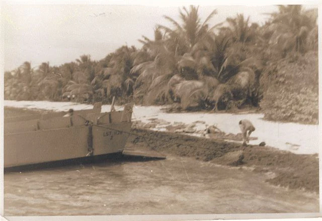 A sepia-toned vintage photograph shows a small landing craft beached on a sandy shore next to palm trees.