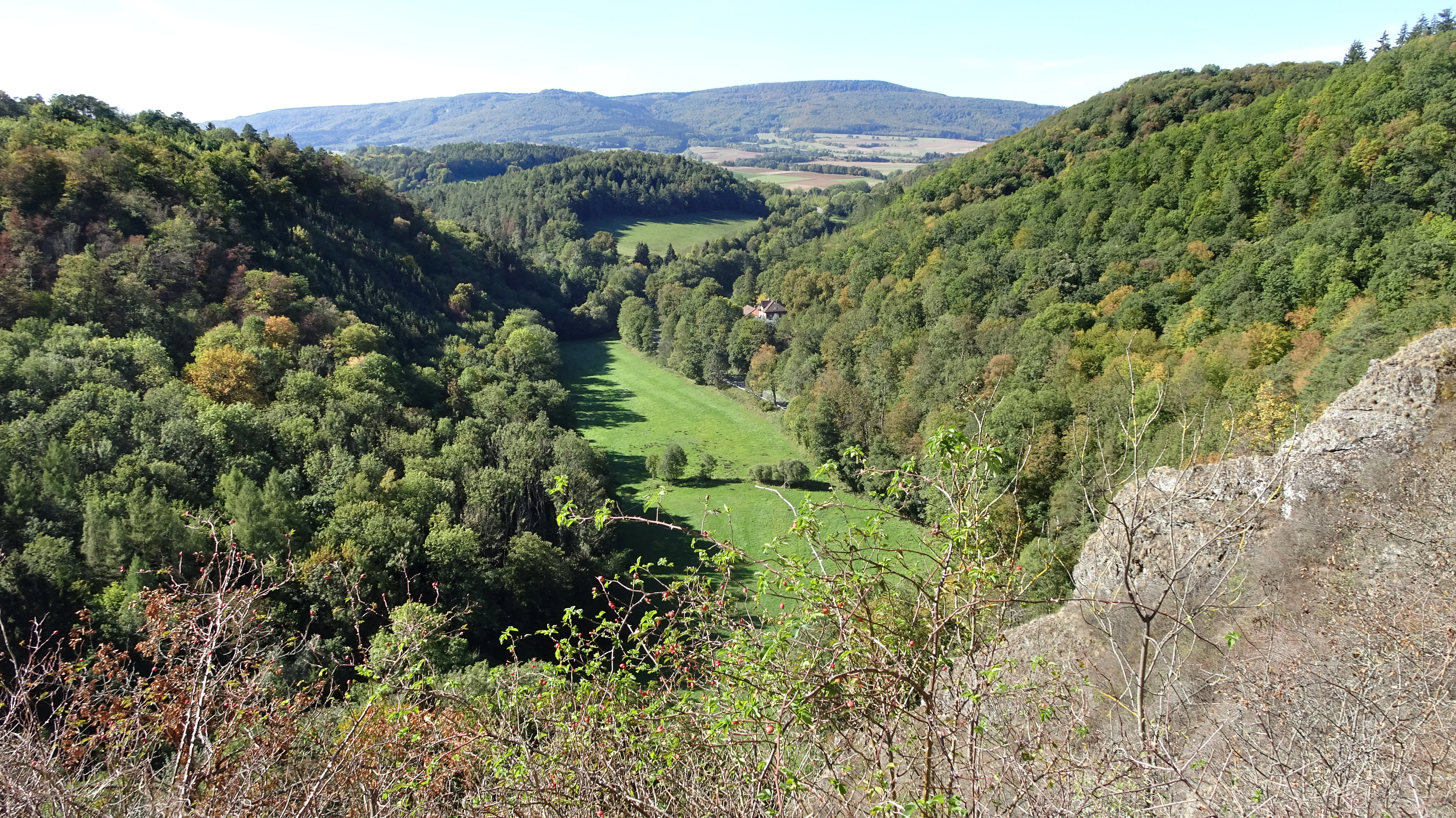 Una vista panorámica en ángulo elevado de un valle profundo y verde rodeado de colinas boscosas, con acantilados rocosos visibles en primer plano a la derecha.