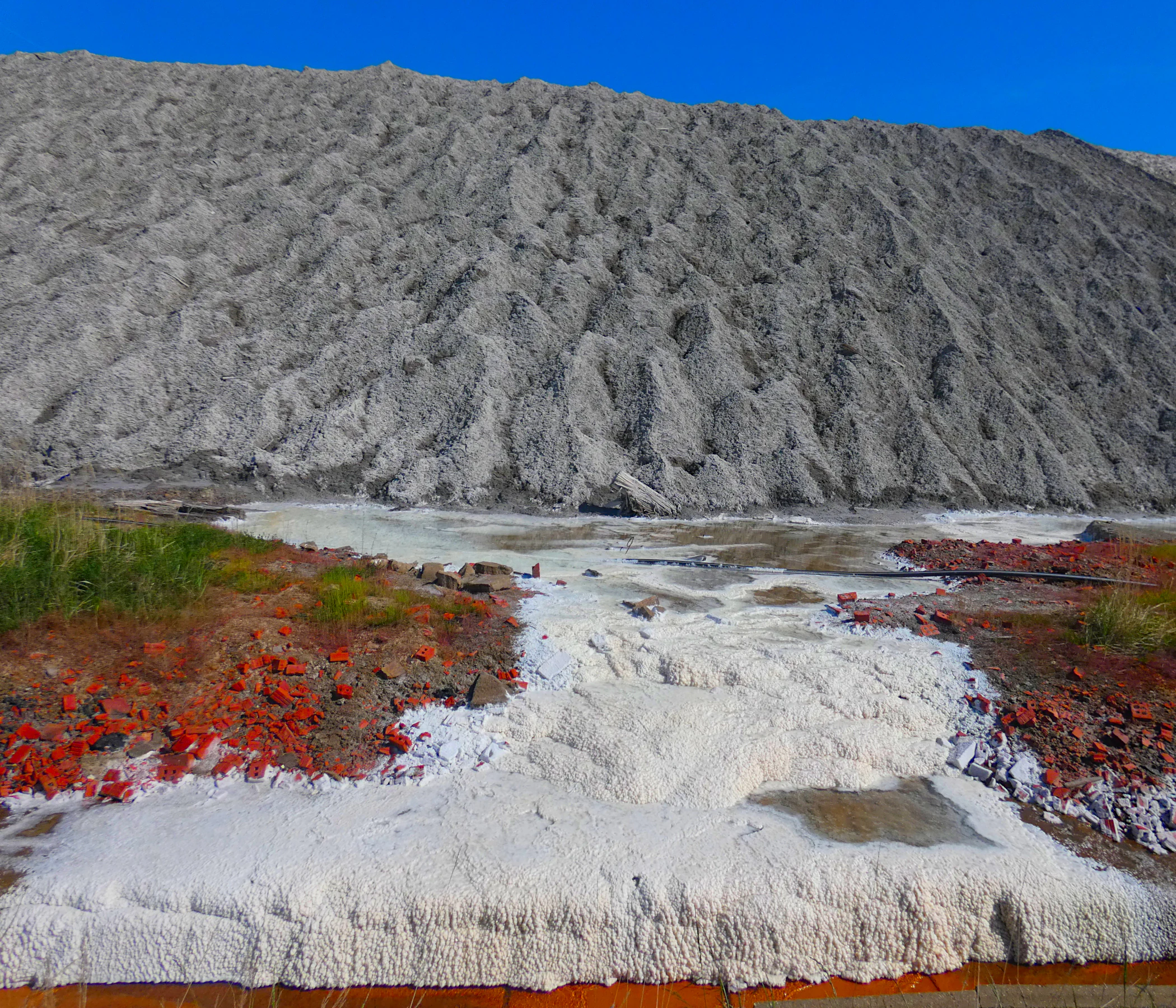 A wide, textured grey waste pile rises behind a foreground of salt deposits and red-colored vegetation.