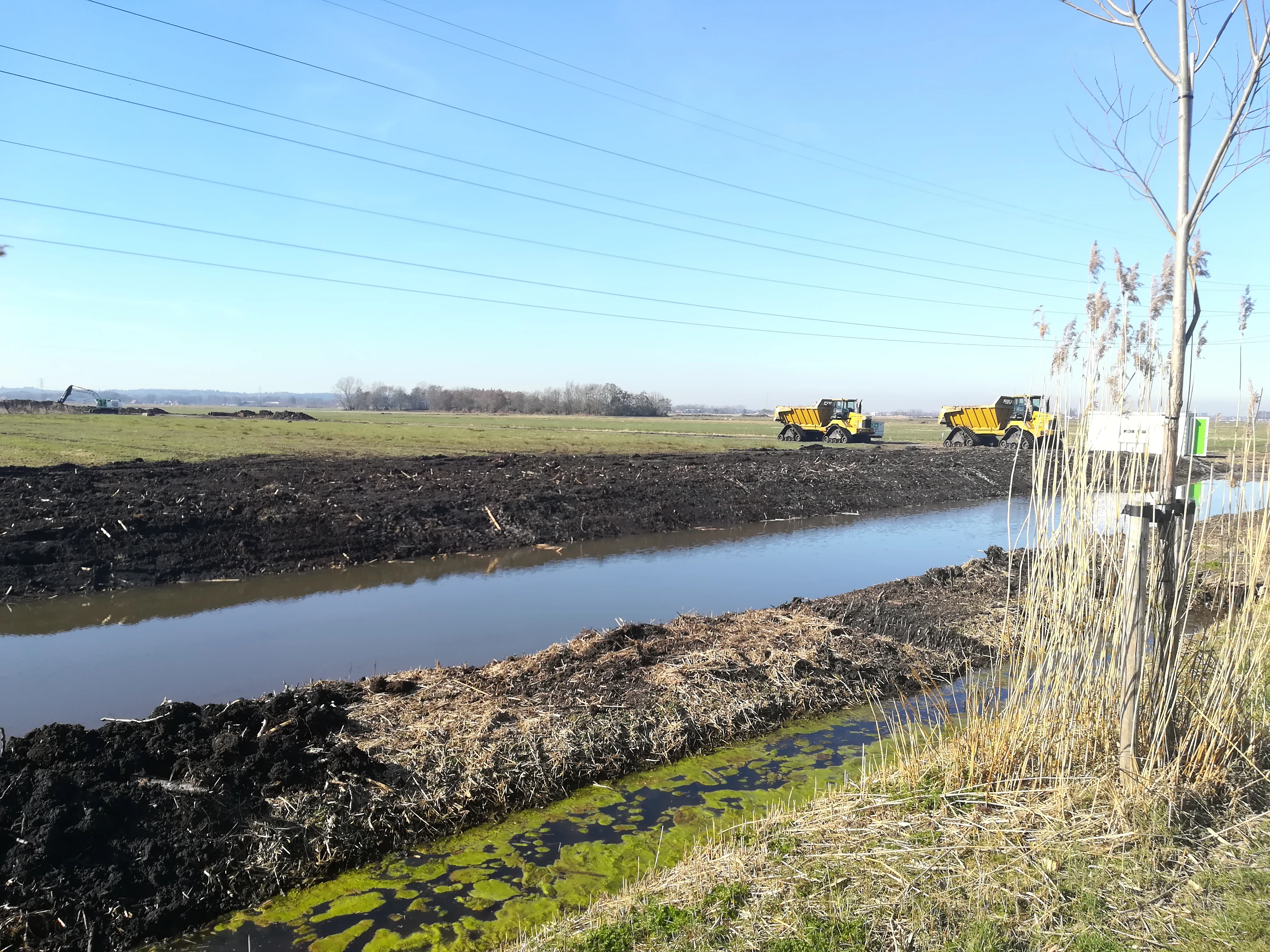 Una vista amplia de un paisaje neerlandés de humedales con un canal estrecho en primer plano, orillas de tierra excavada y vehículos de construcción amarillos operando a lo lejos bajo un cielo azul despejado.