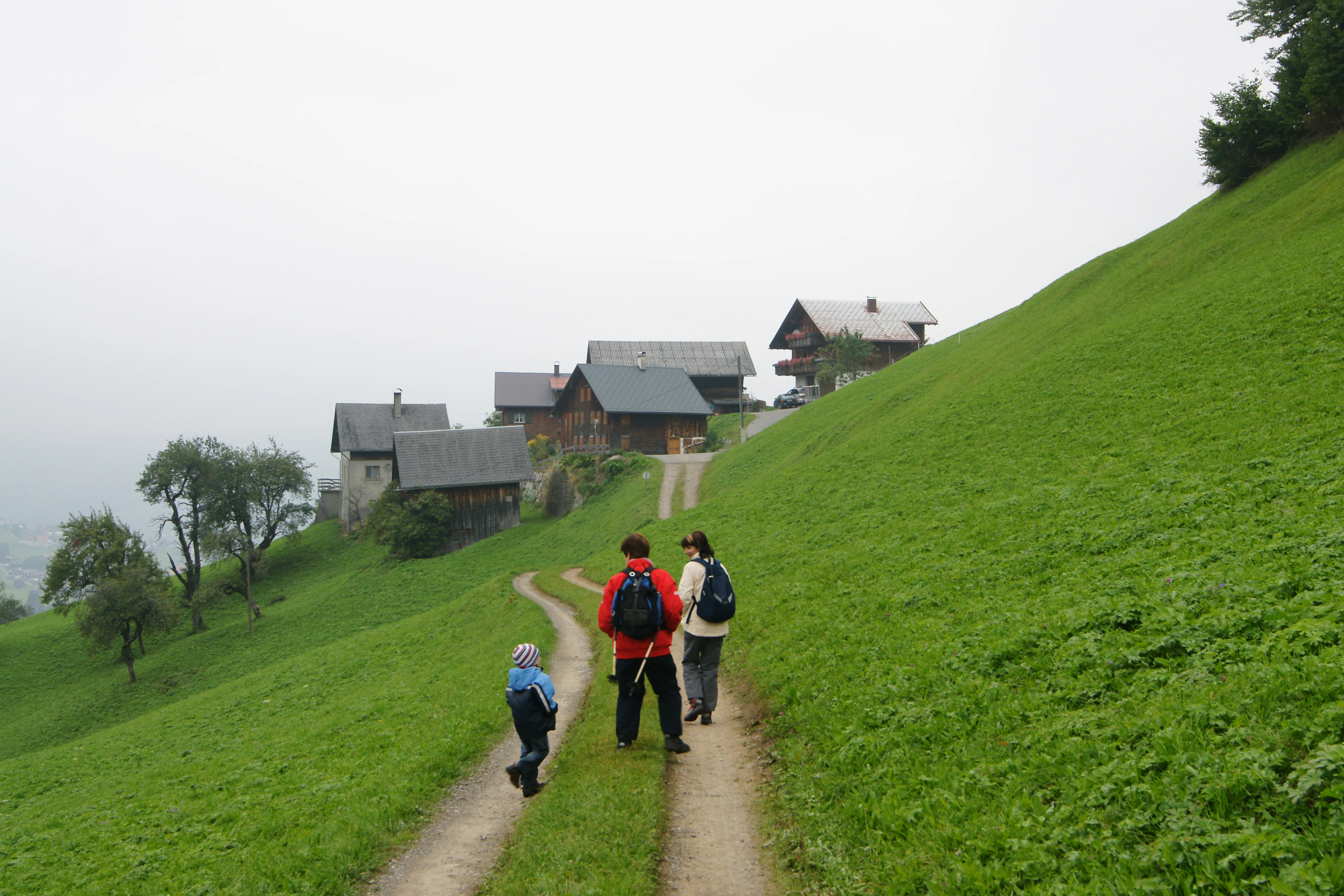 Three hikers walking away from the camera on a dirt trail leading up a green hillside toward a cluster of wooden farmhouses under an overcast sky.