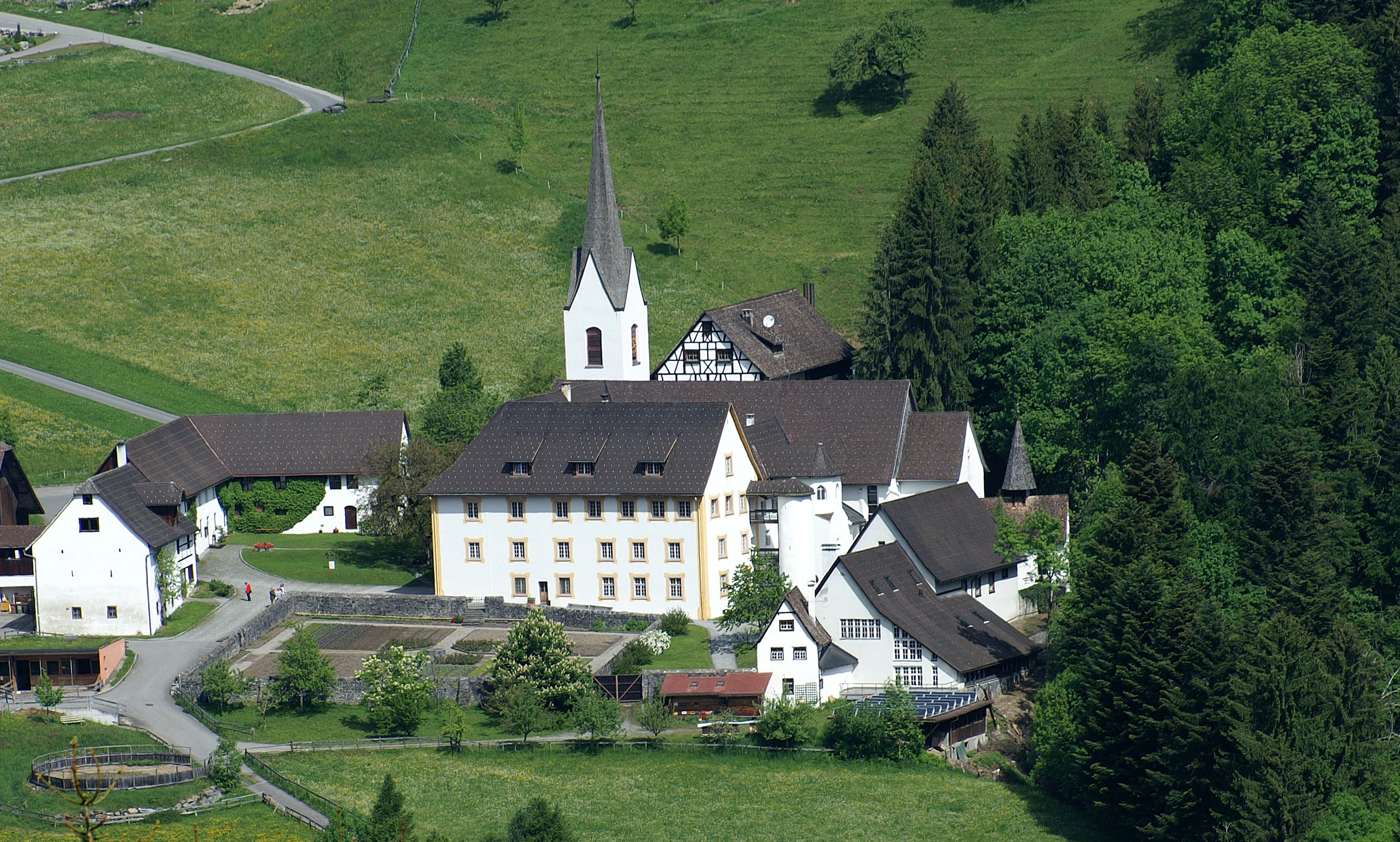 High-angle view of a white priory complex with a tall church spire, surrounded by green meadows and coniferous forests.